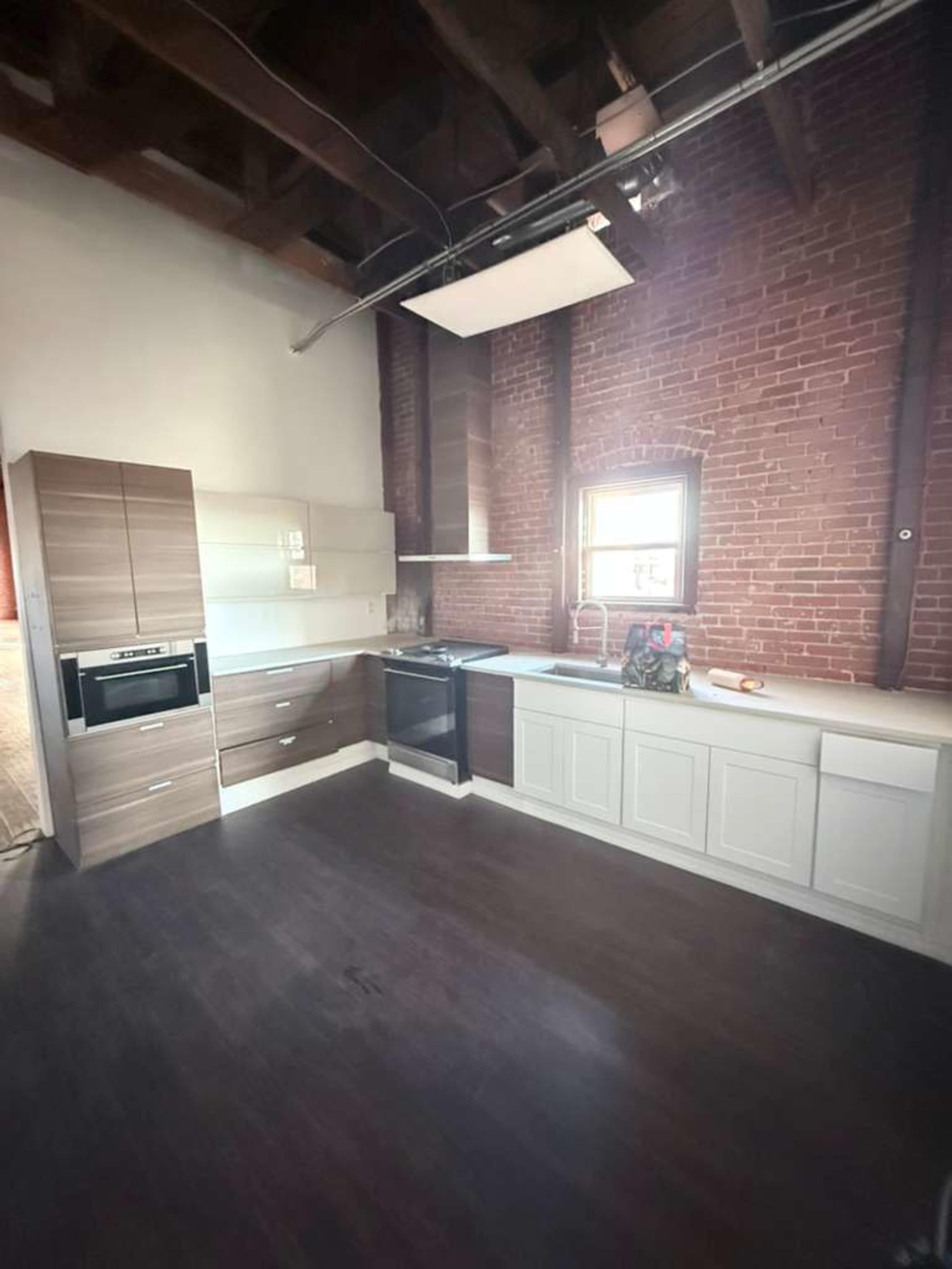 The image shows a modern kitchen with a combination of light and dark cabinetry against a backdrop of exposed brick walls and wooden beams.