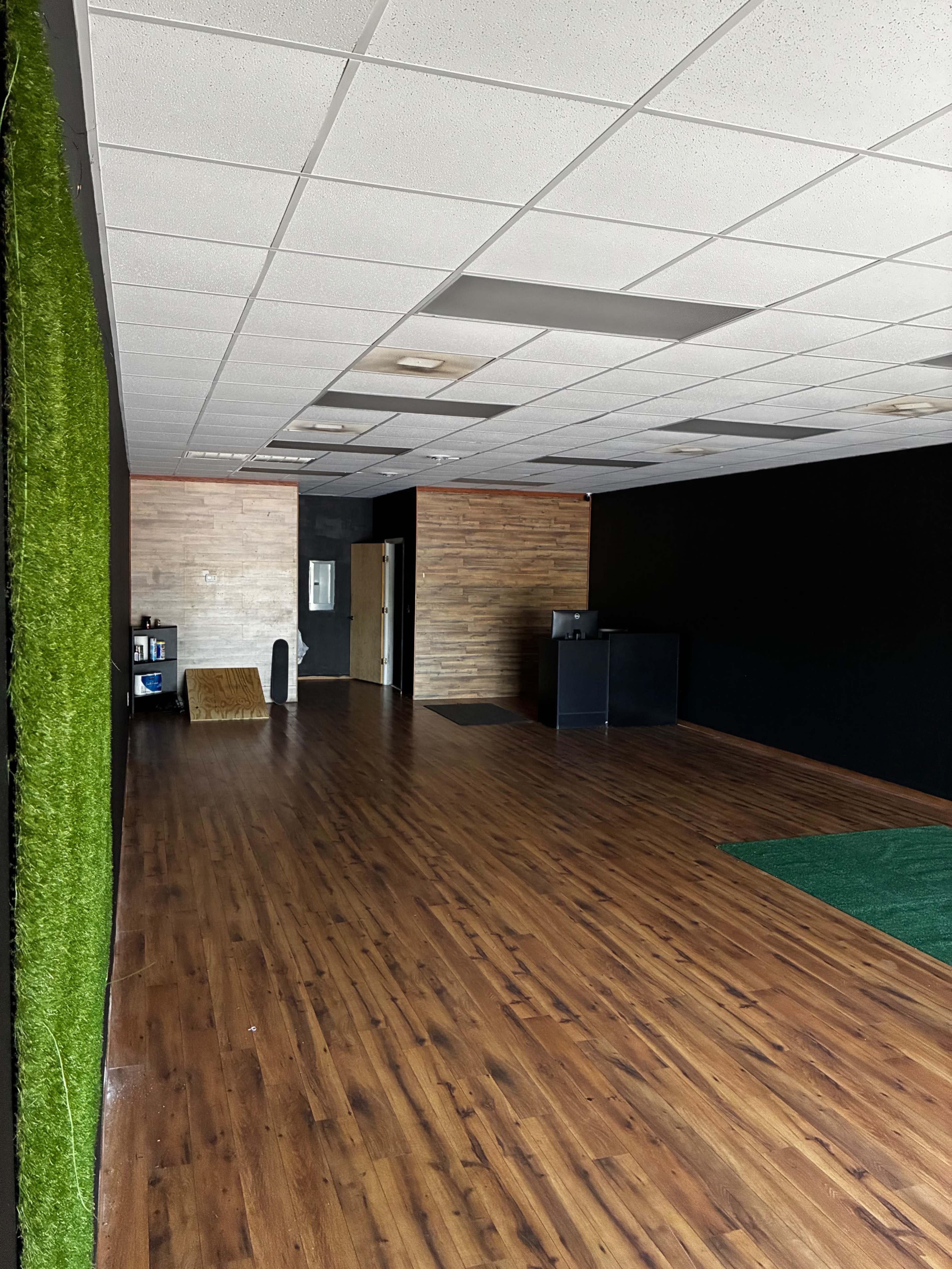 The image shows an empty interior space featuring a wooden floor, a black accent wall, and a section of artificial grass on the right.