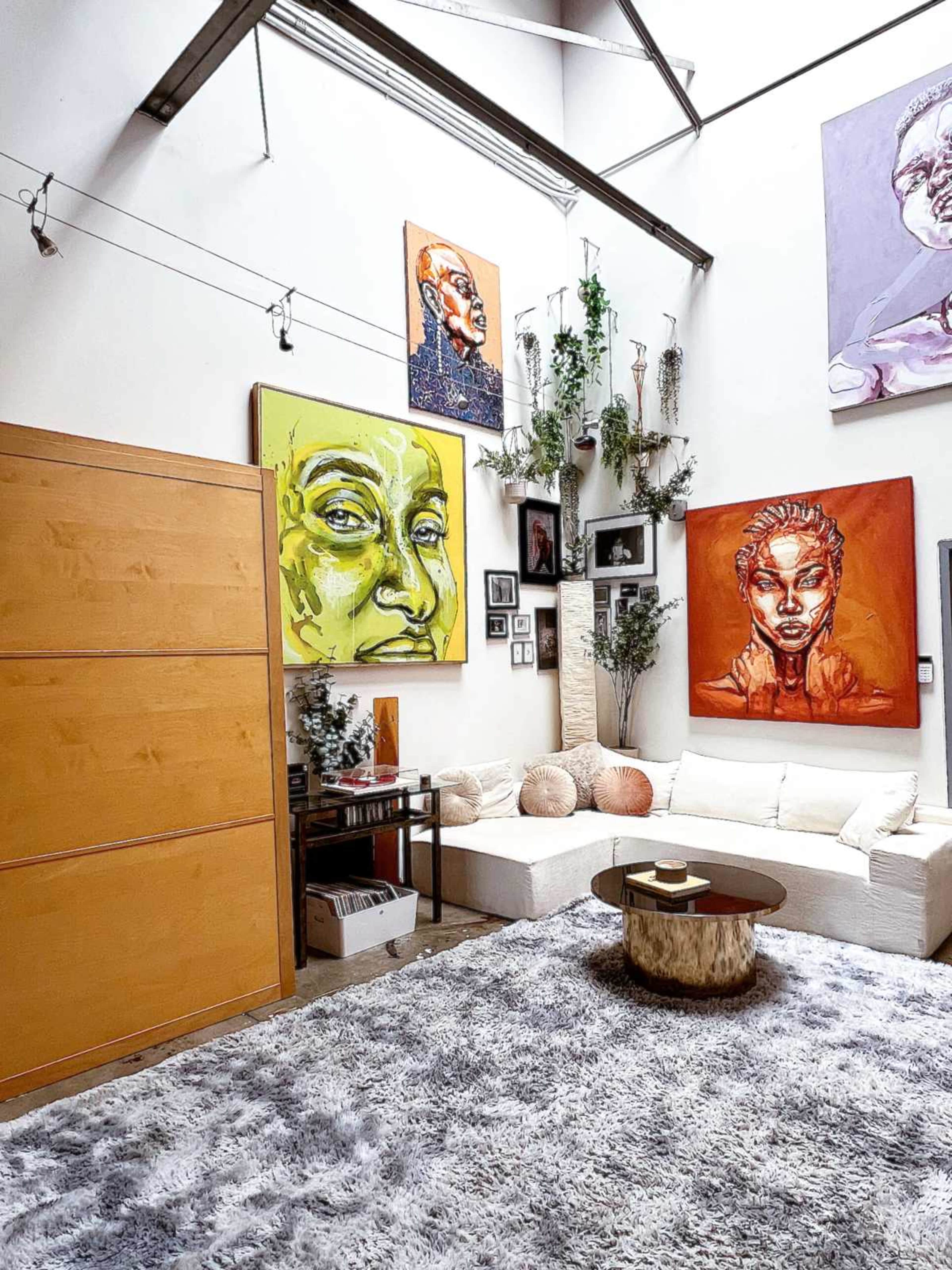 The image shows a stylish living space with large colorful portraits on the walls, a white sectional sofa, a round coffee table, and a plush gray rug.