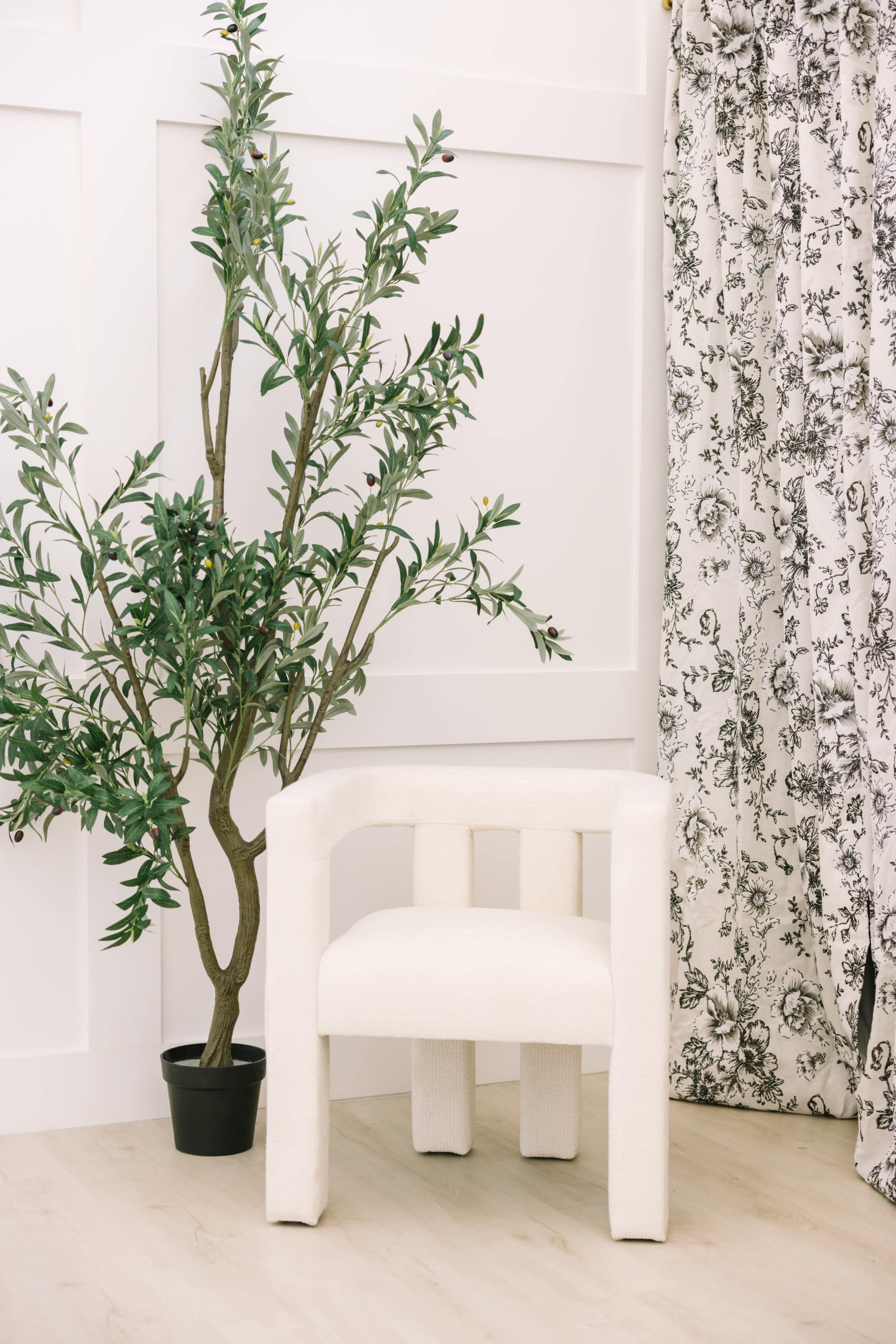 A white upholstered chair is positioned next to a potted plant, in front of a floral-patterned curtain against a white wall.