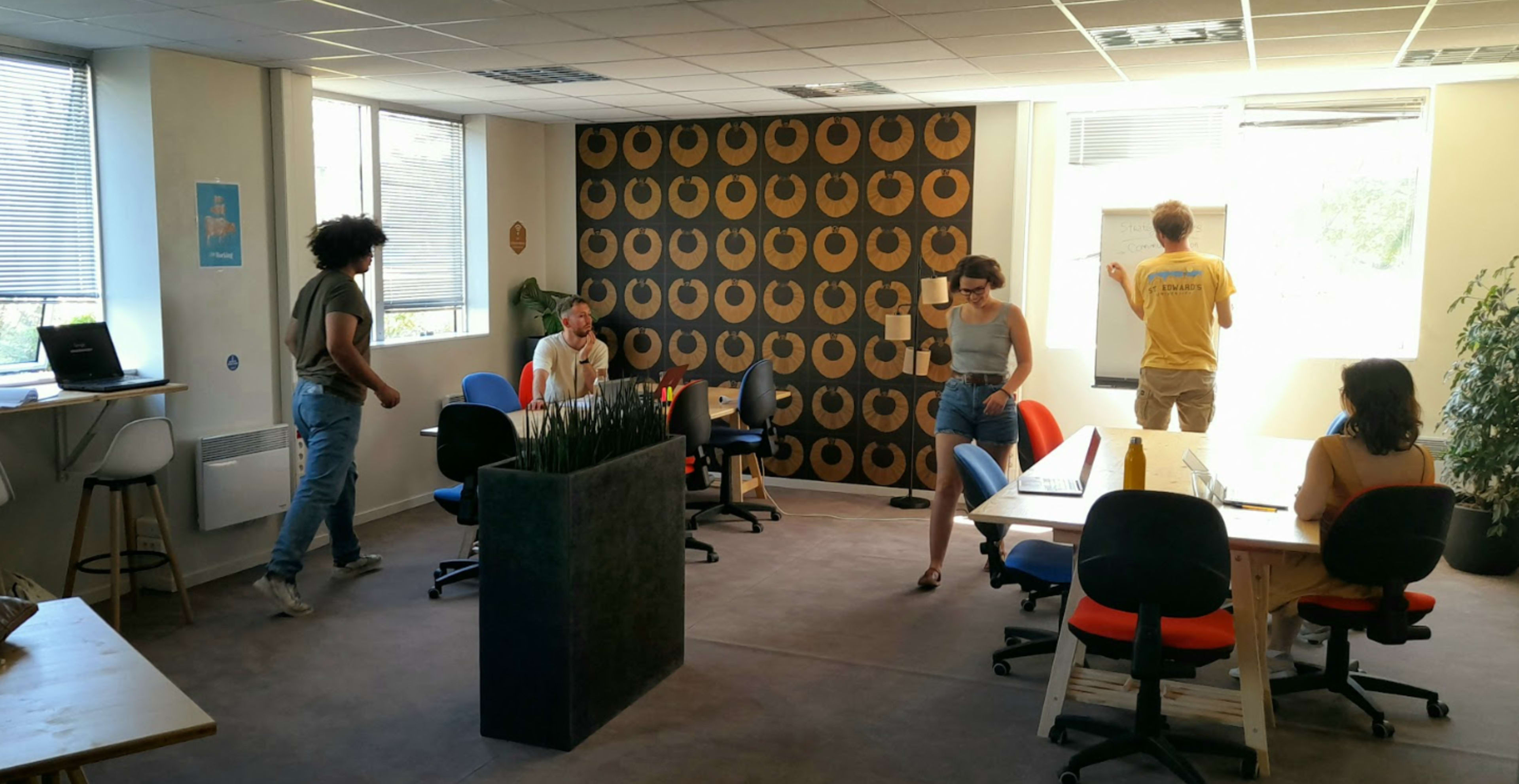 A group of five people work in a modern office space with colorful chairs and a feature wall adorned with a circular pattern.