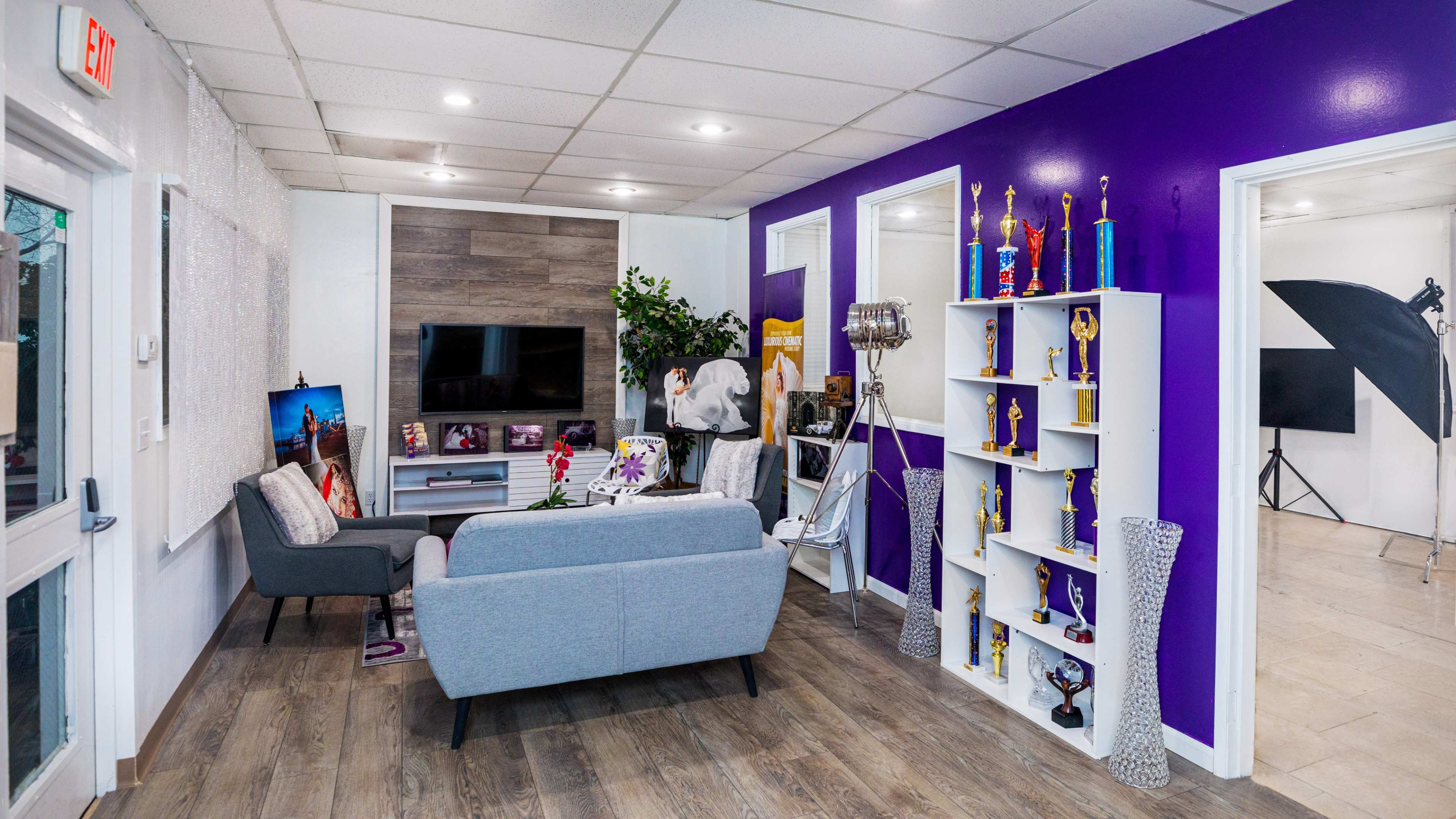 A modern interior space featuring a gray sofa, a television, a shelf with trophies, and purple walls, with an area for photography in the background.
