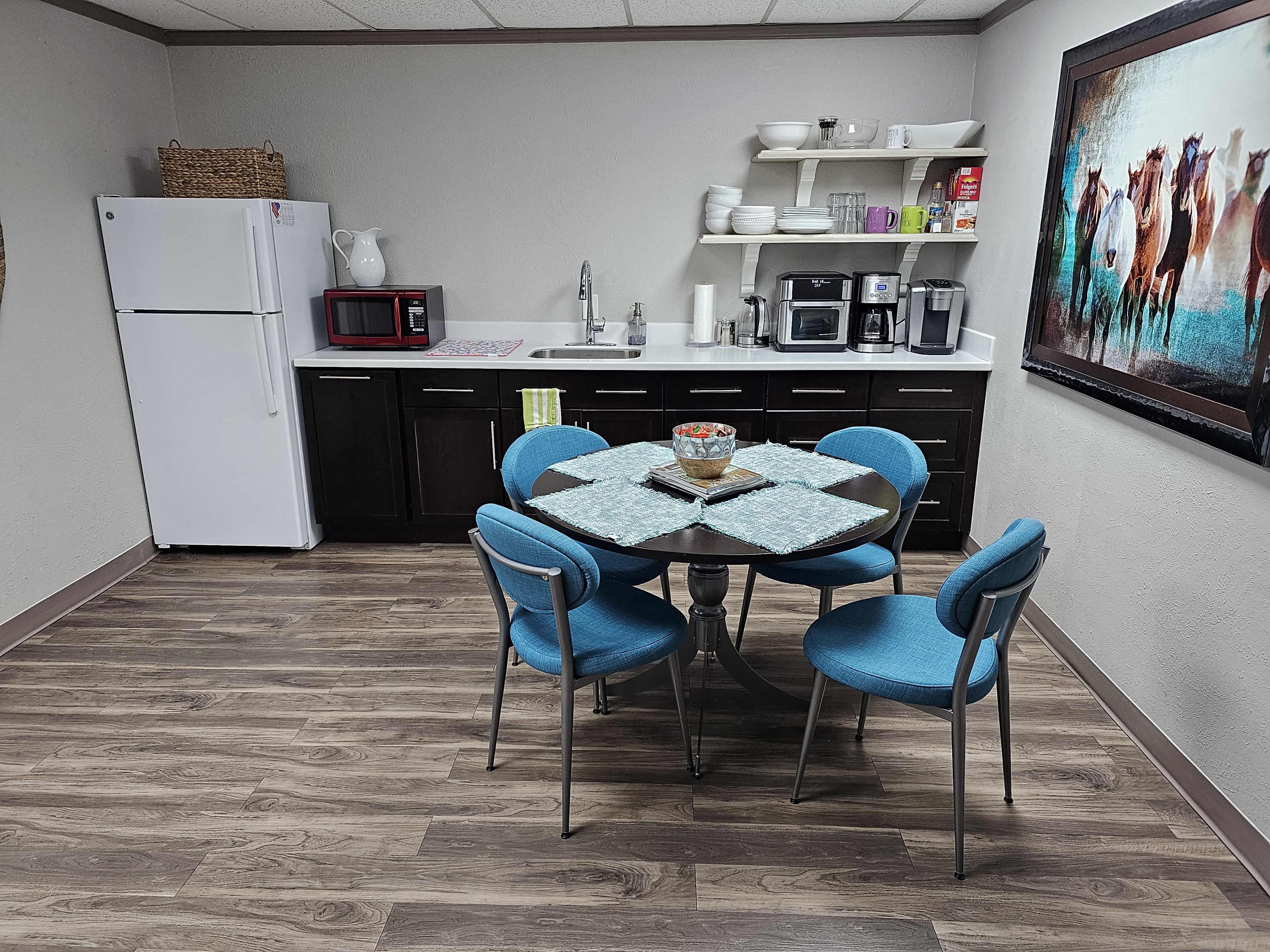 The image shows a small kitchen area with a round table and four blue chairs, along with a refrigerator, microwave, sink, and coffee maker.