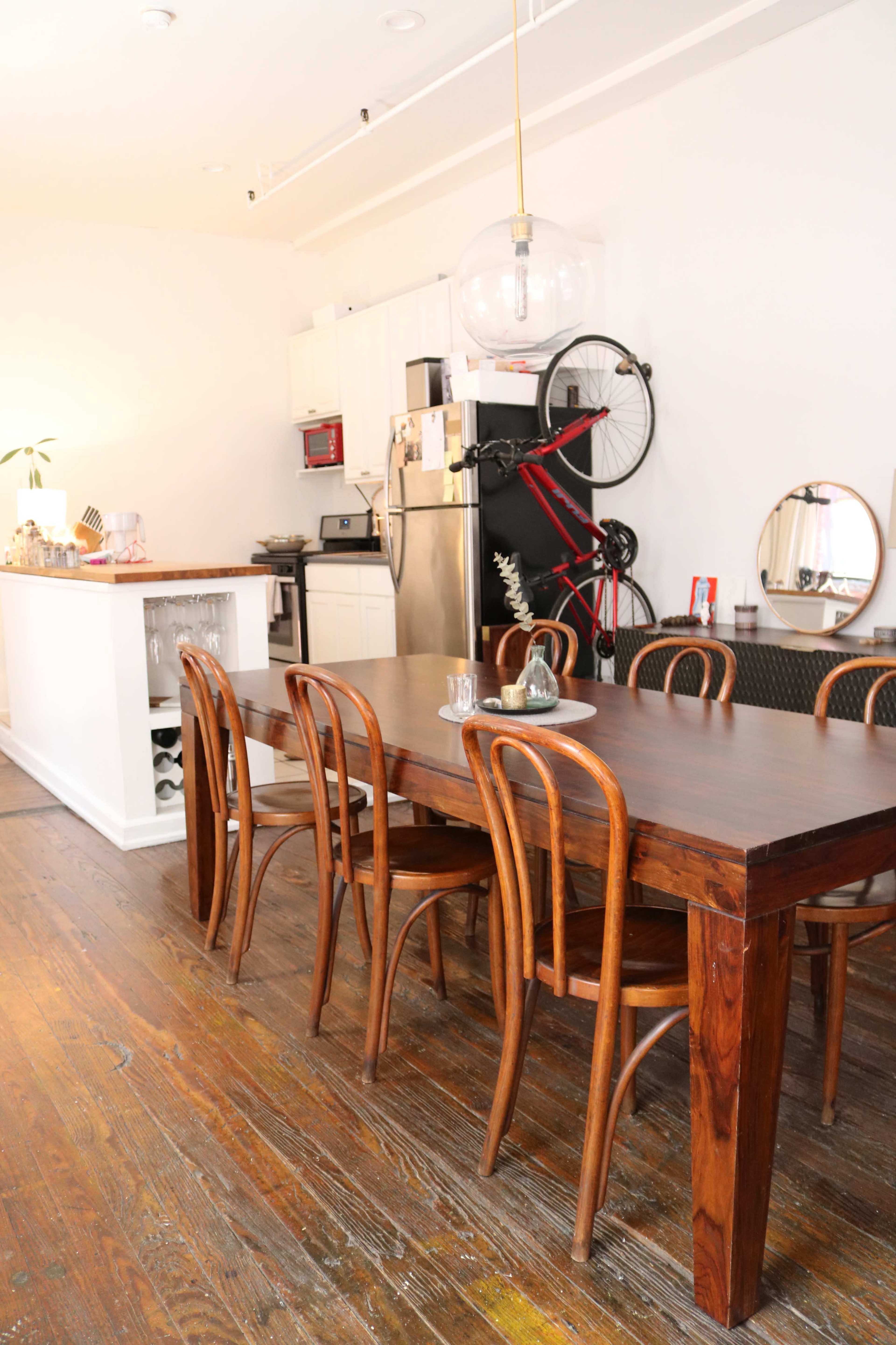 A long wooden dining table with several chairs is positioned in a room featuring a kitchen area with modern appliances and a bicycle hanging on the wall.