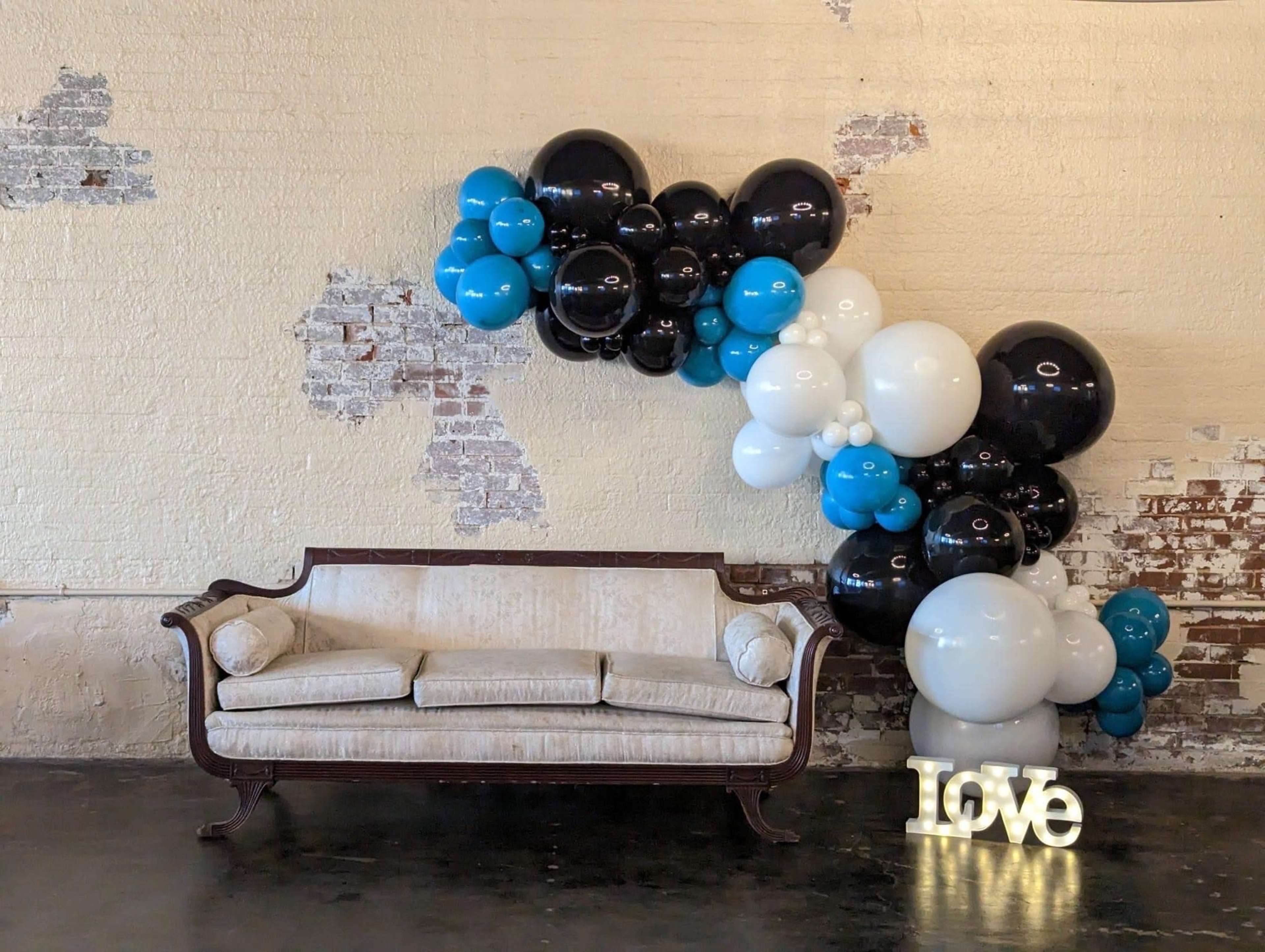 A vintage sofa is positioned against a textured wall adorned with a cluster of black, white, and blue balloons, alongside a decorative light-up "Love" sign.