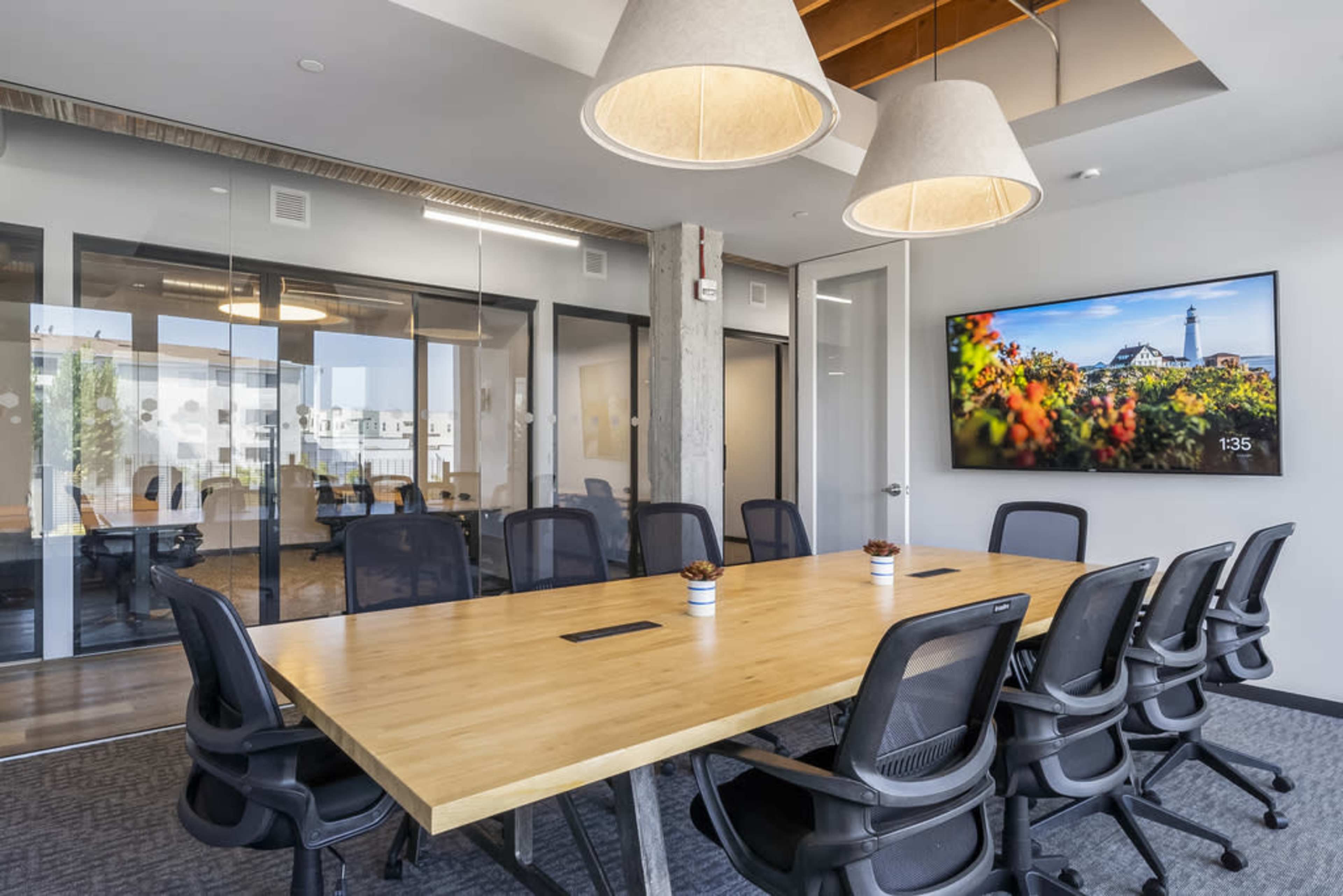 A modern conference room features a long wooden table surrounded by black ergonomic chairs, with a large screen displaying an image of a lighthouse.