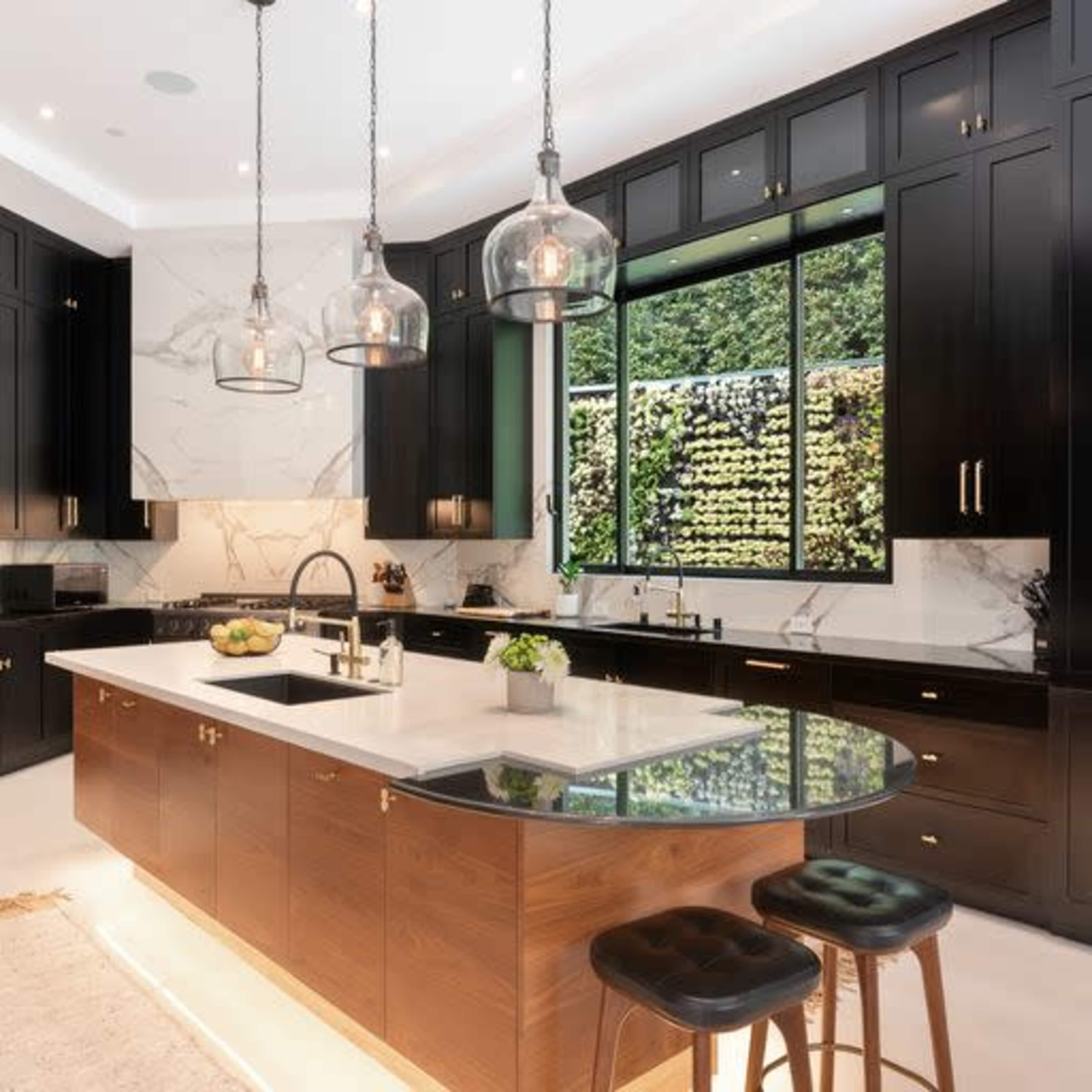 A modern kitchen featuring dark cabinetry, marble countertops, and pendant lighting, with a large window overlooking greenery.