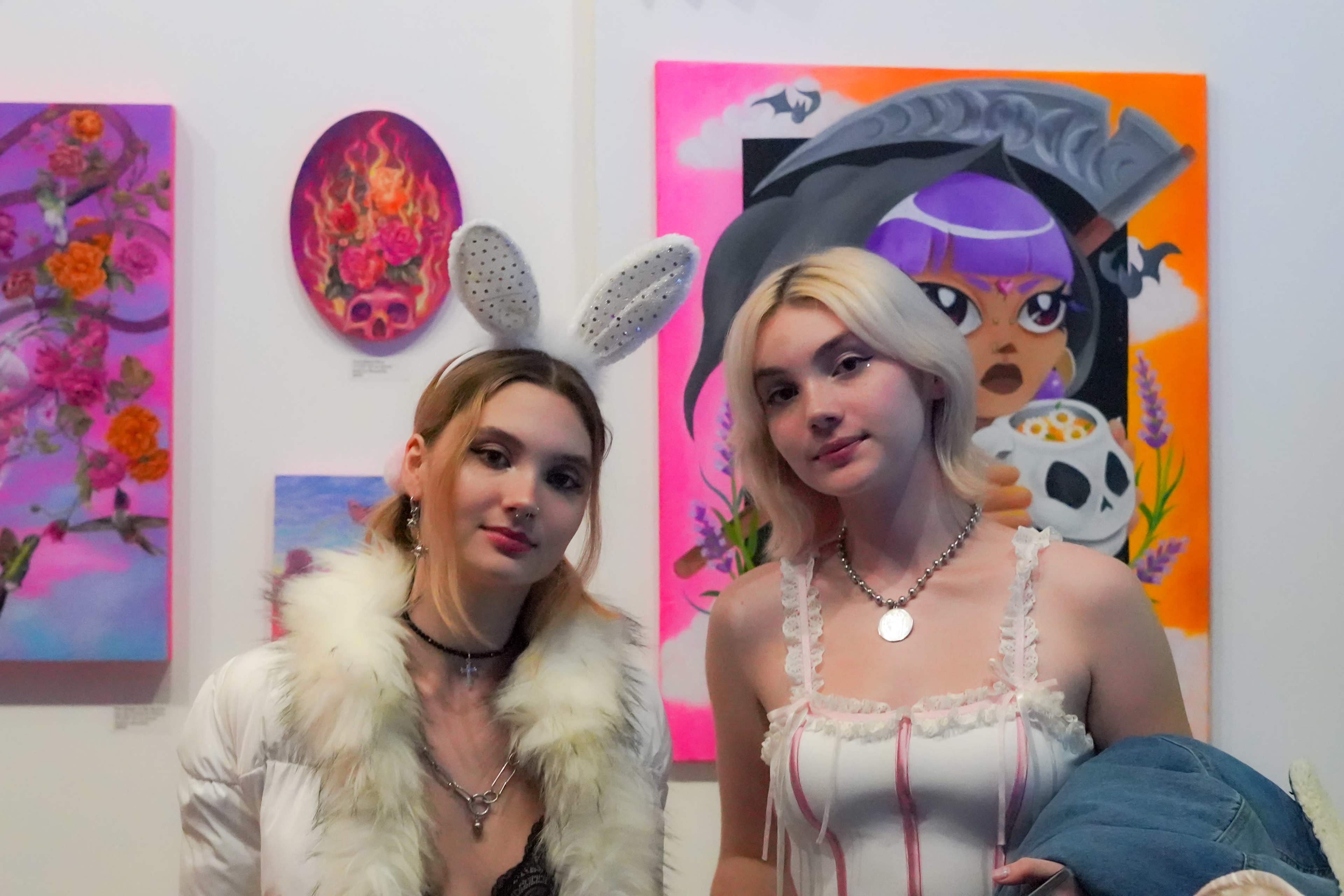 Two young women pose for a photo in an art gallery with colorful paintings on the wall behind them.