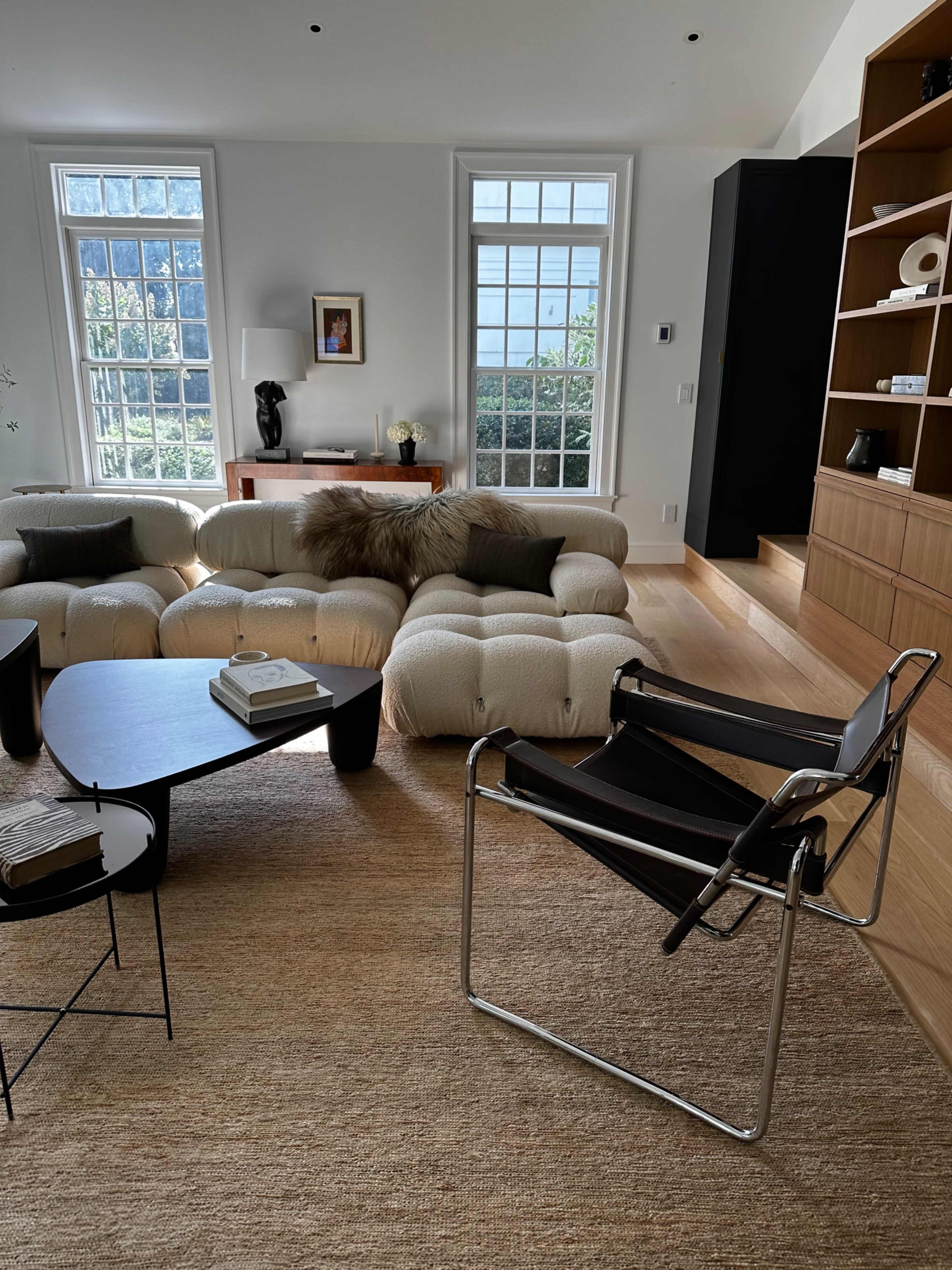 The living room features a plush, light-colored sofa, a black coffee table, and a sleek, modern chair, with large windows allowing natural light to fill the space.