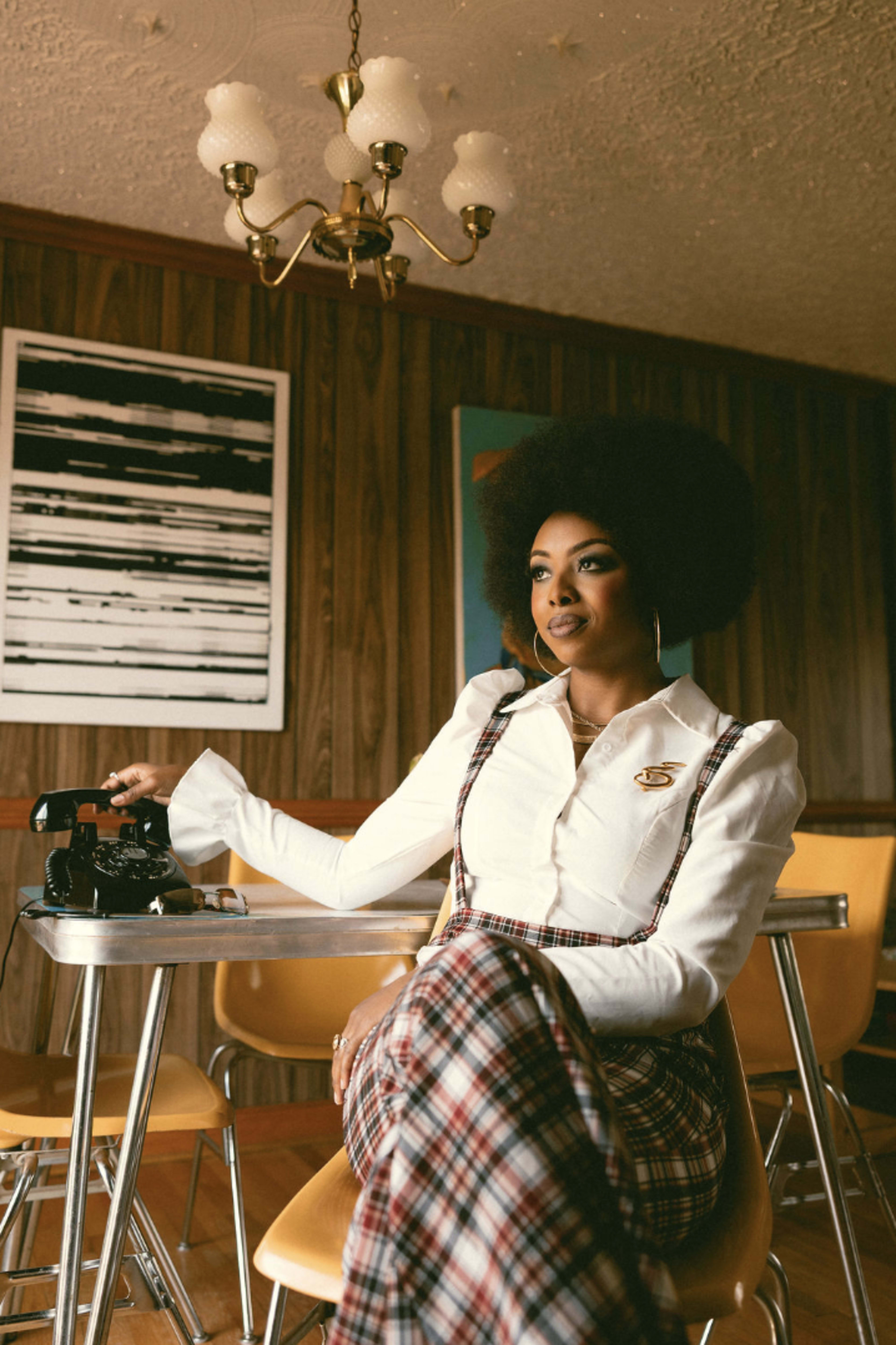 A woman with an afro hairstyle sits at a table in a retro-style room with wooden paneling, featuring a vintage telephone and a chandelier above.