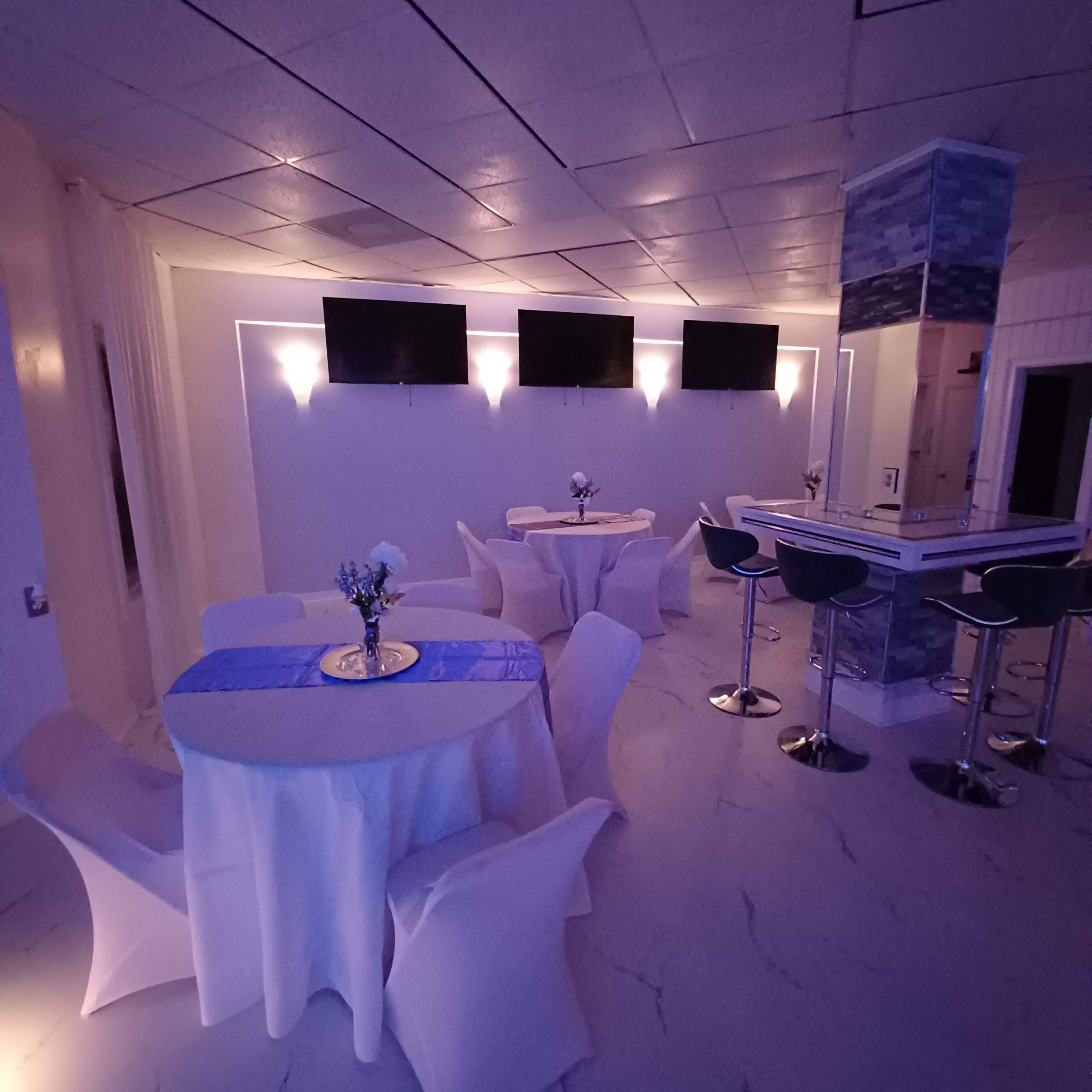 A modern interior space with white tables, chairs, and several television screens mounted on the walls.