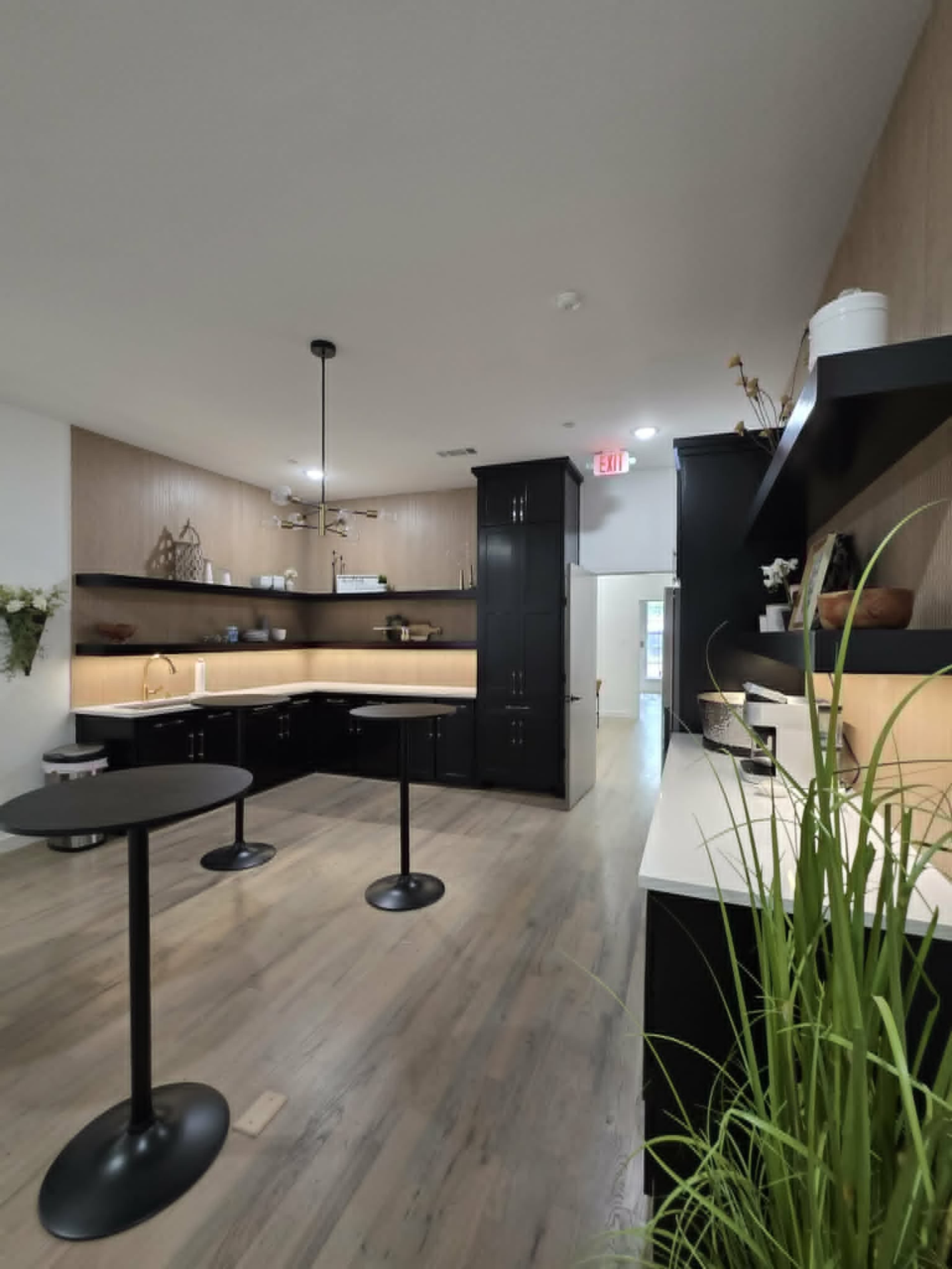 The image shows a modern kitchen space featuring black cabinetry, countertops, and two tall, round tables, with a light wood accent wall and a doorway leading to another room.