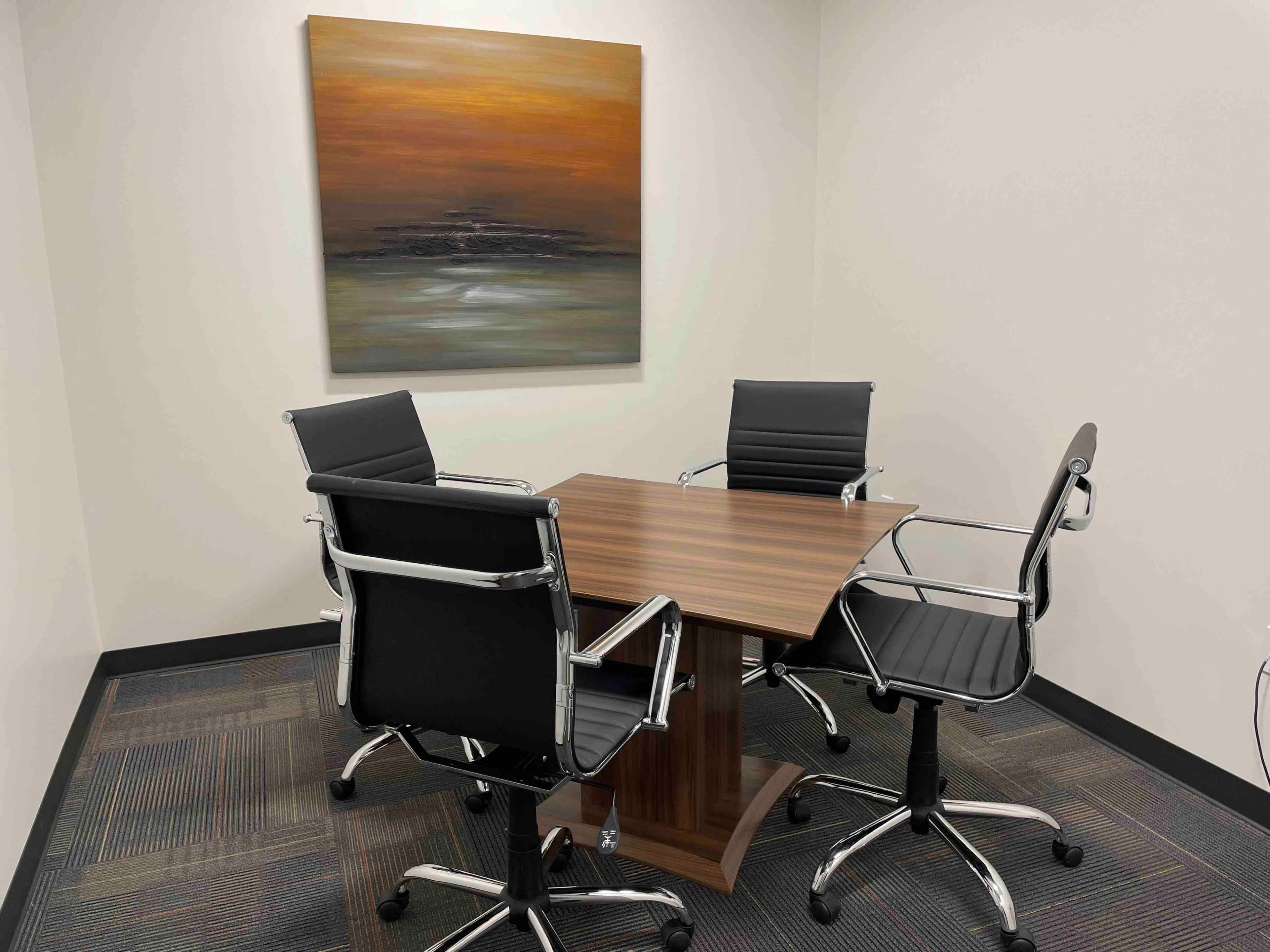 A small conference room features a wooden table surrounded by four black chairs and an abstract painting on the wall.