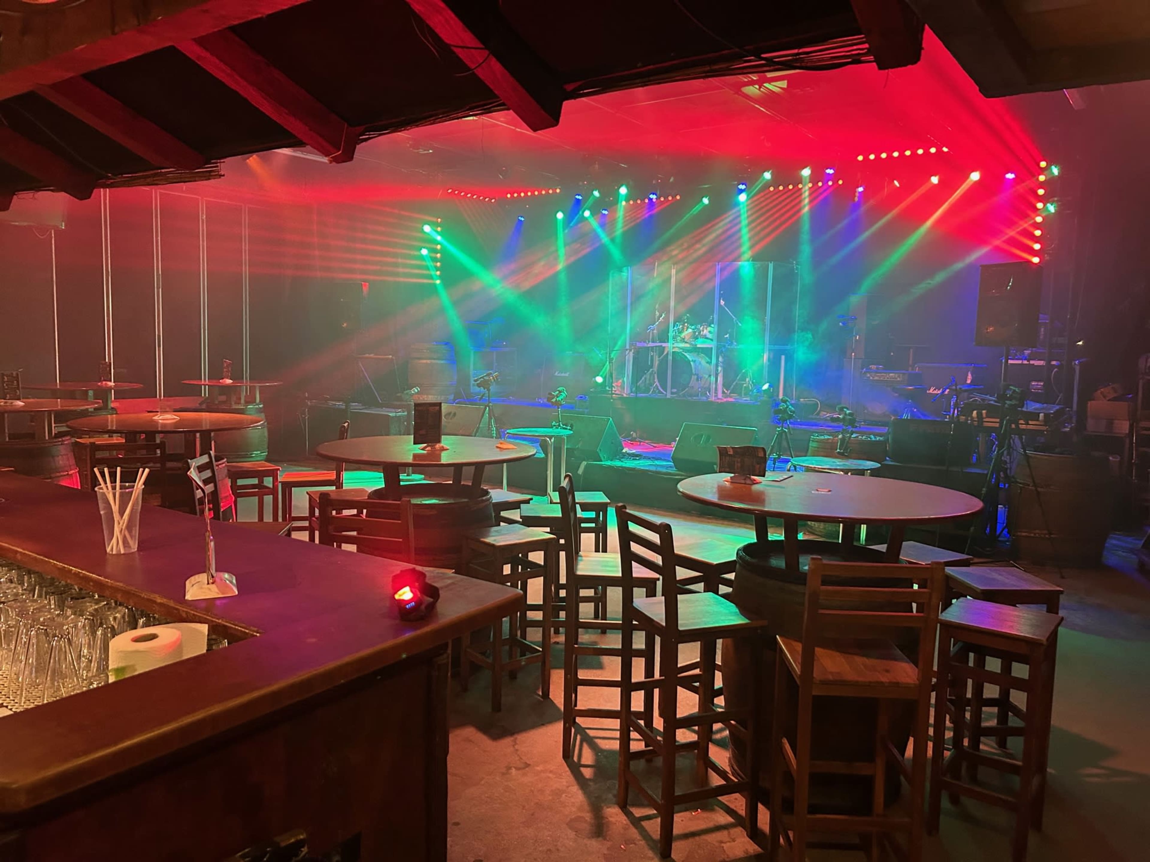 A dimly lit live music venue with tables and chairs arranged in front of a stage, illuminated by colorful stage lights and smoke.
