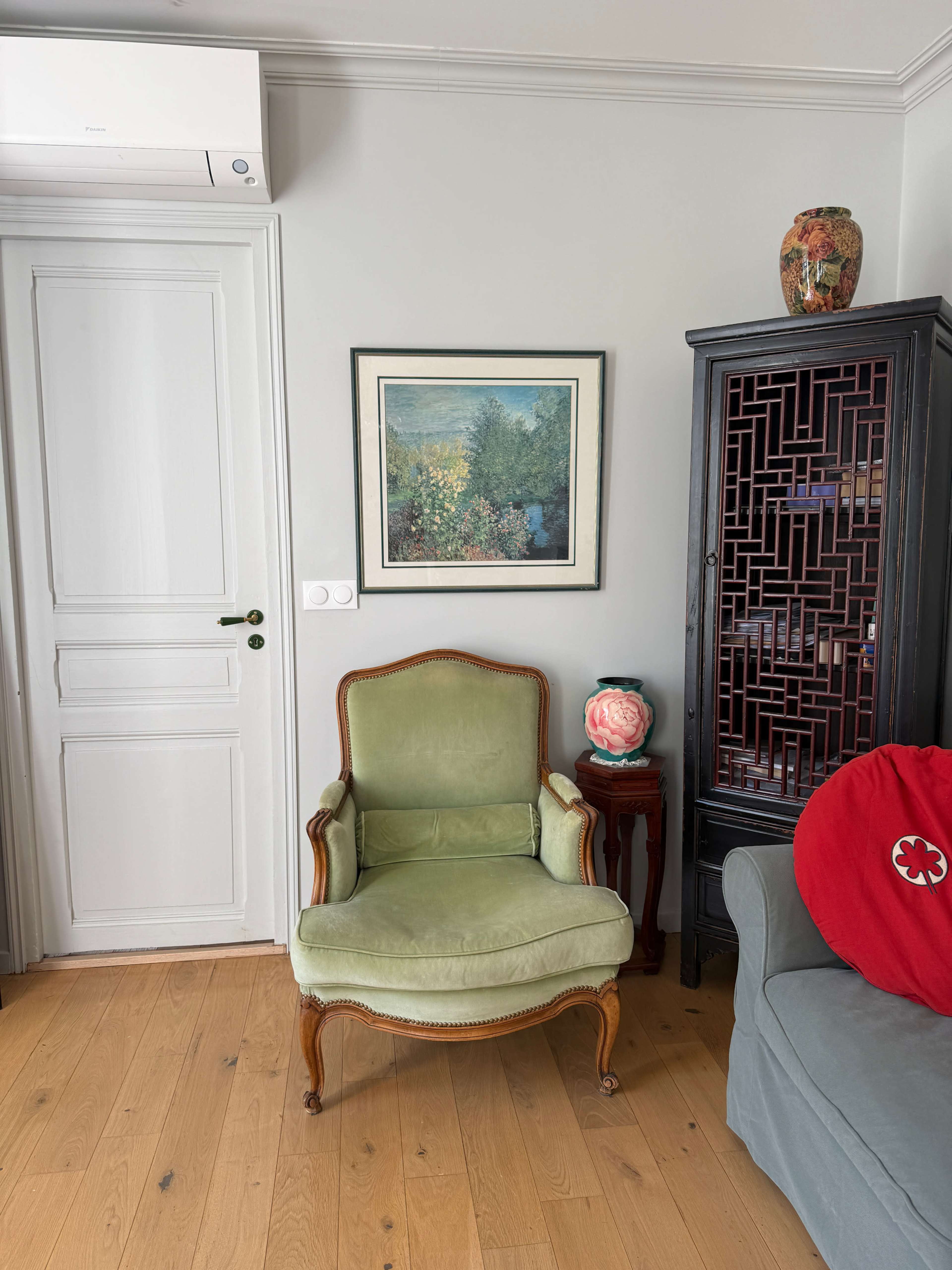 The image shows a green upholstered armchair beside a dark wooden cabinet, with a floral painting hanging on the wall and a vase on top of the cabinet.