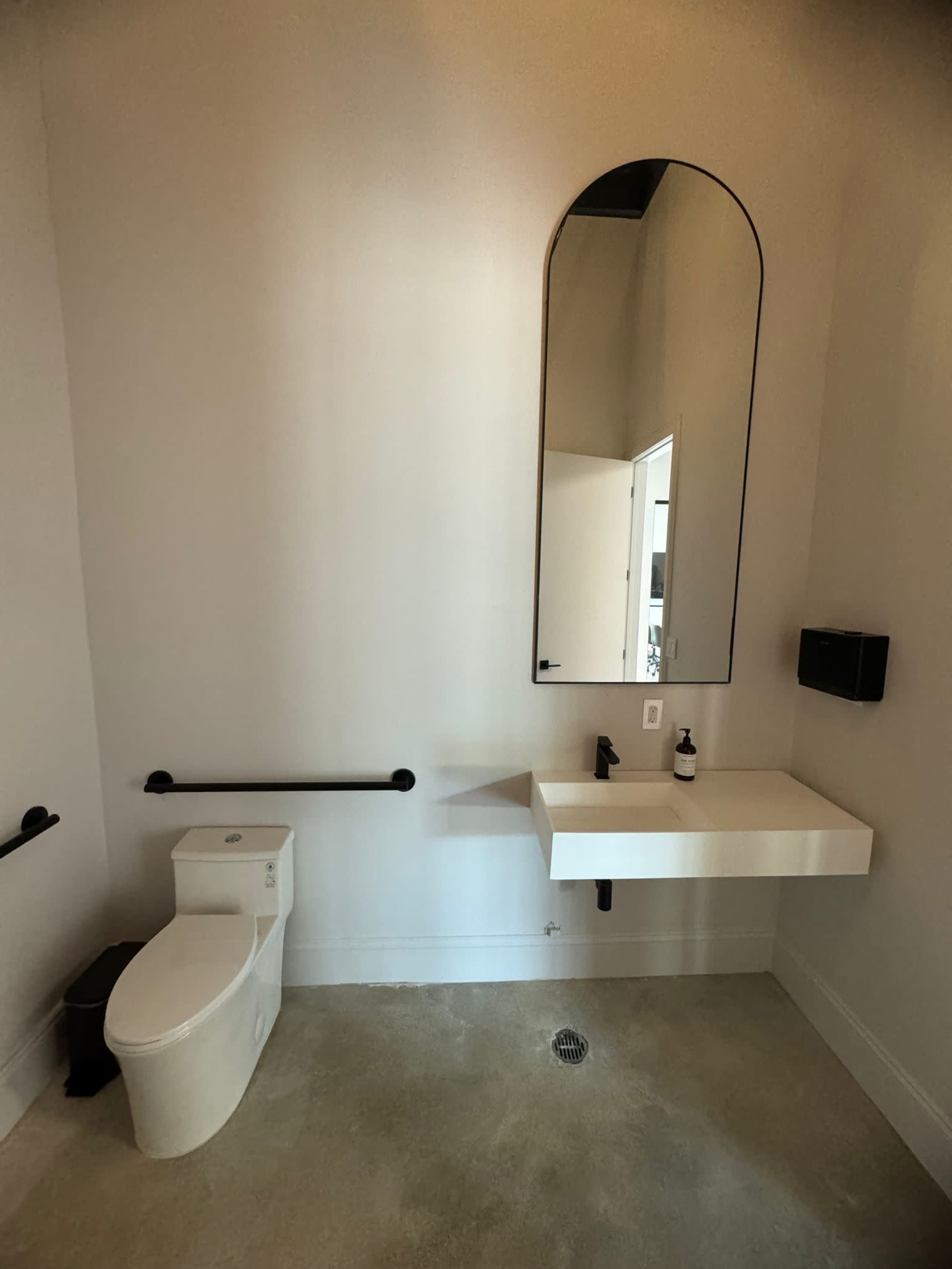 A modern bathroom featuring a wall-mounted sink, a large mirror, a toilet, and grab bars, with minimalistic decor and a concrete floor.