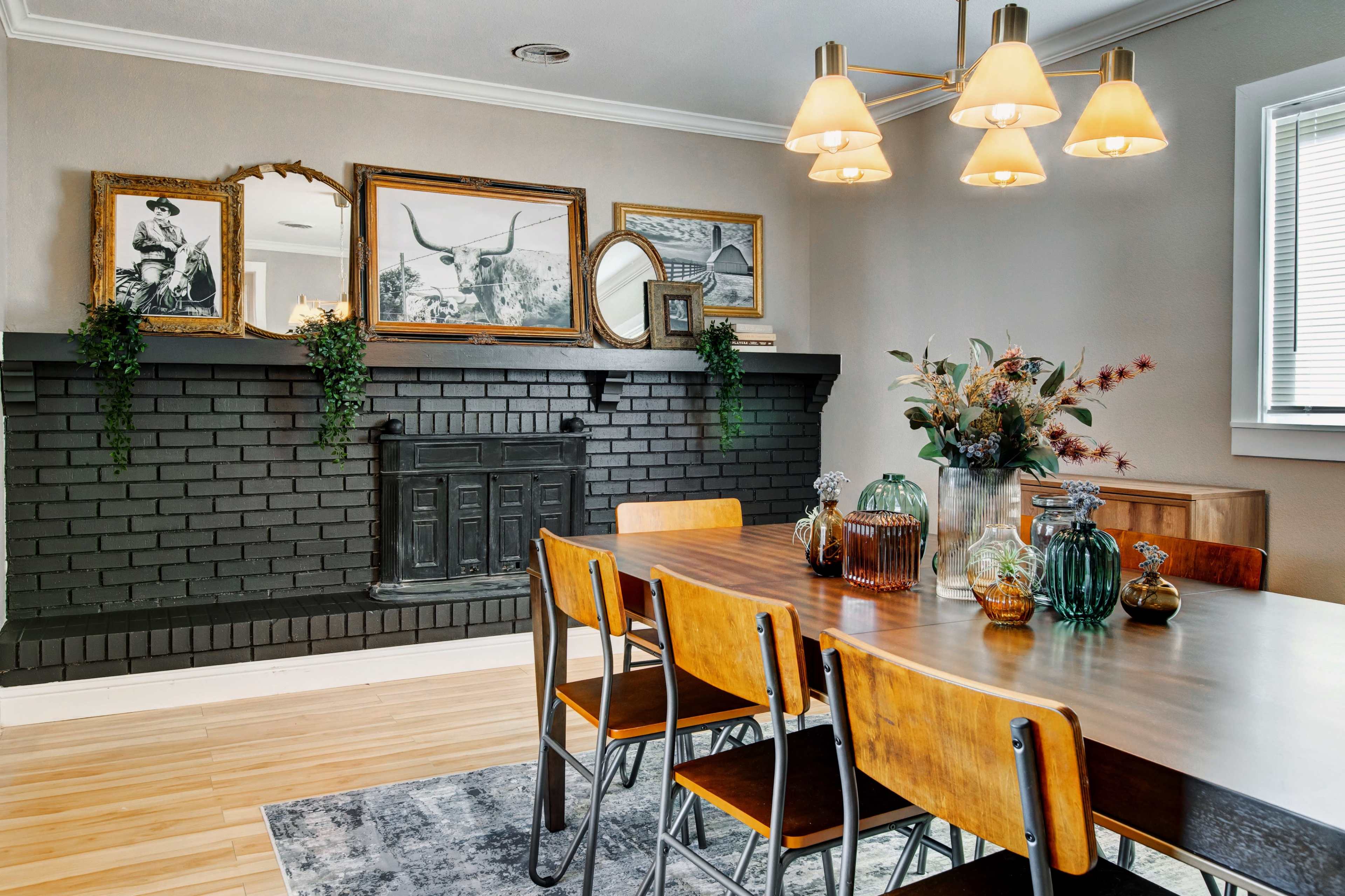 A dining room features a wooden table surrounded by metal chairs, a black brick fireplace with framed artwork above it, and decorative vases on the table.