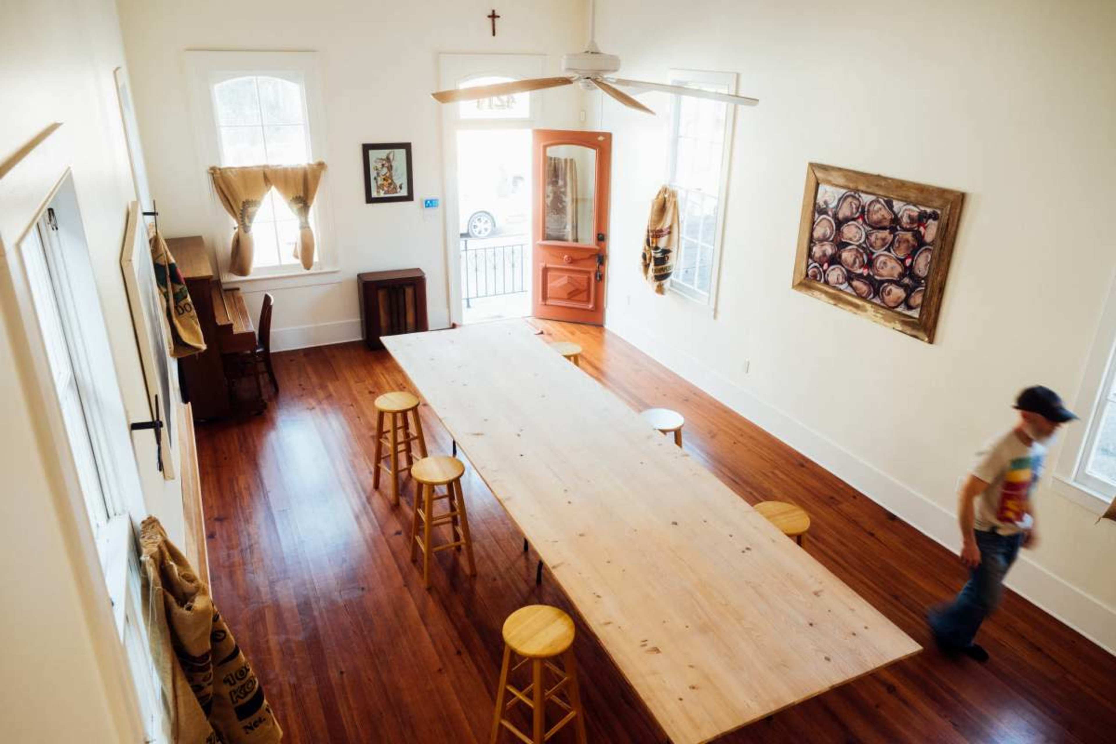 A man walks past a large wooden table surrounded by stools in a bright, minimalist room with wooden flooring and artwork on the walls.