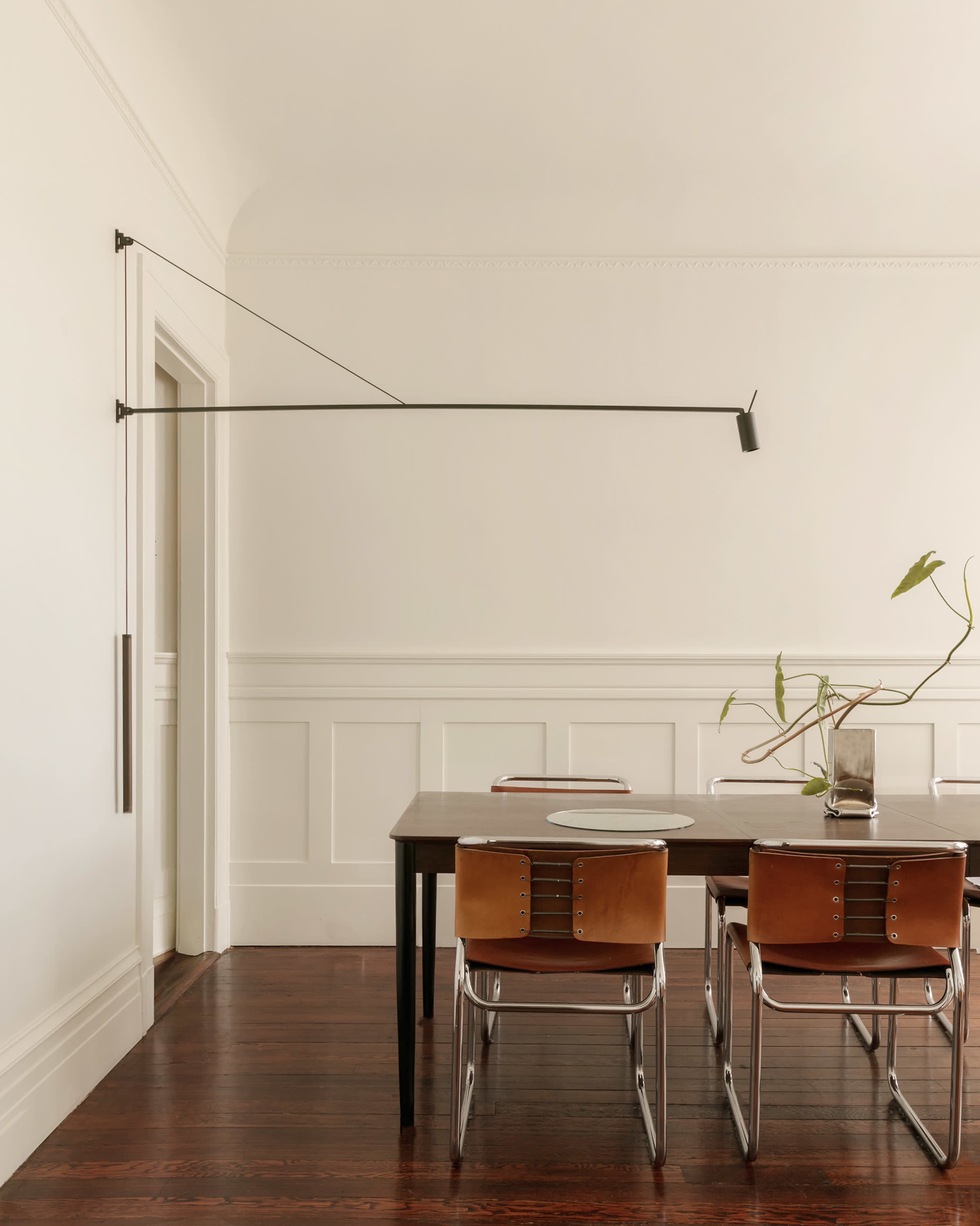 A wooden dining table with leather chairs is set against a plain wall, featuring a wall-mounted lamp extending towards an adjacent doorway.