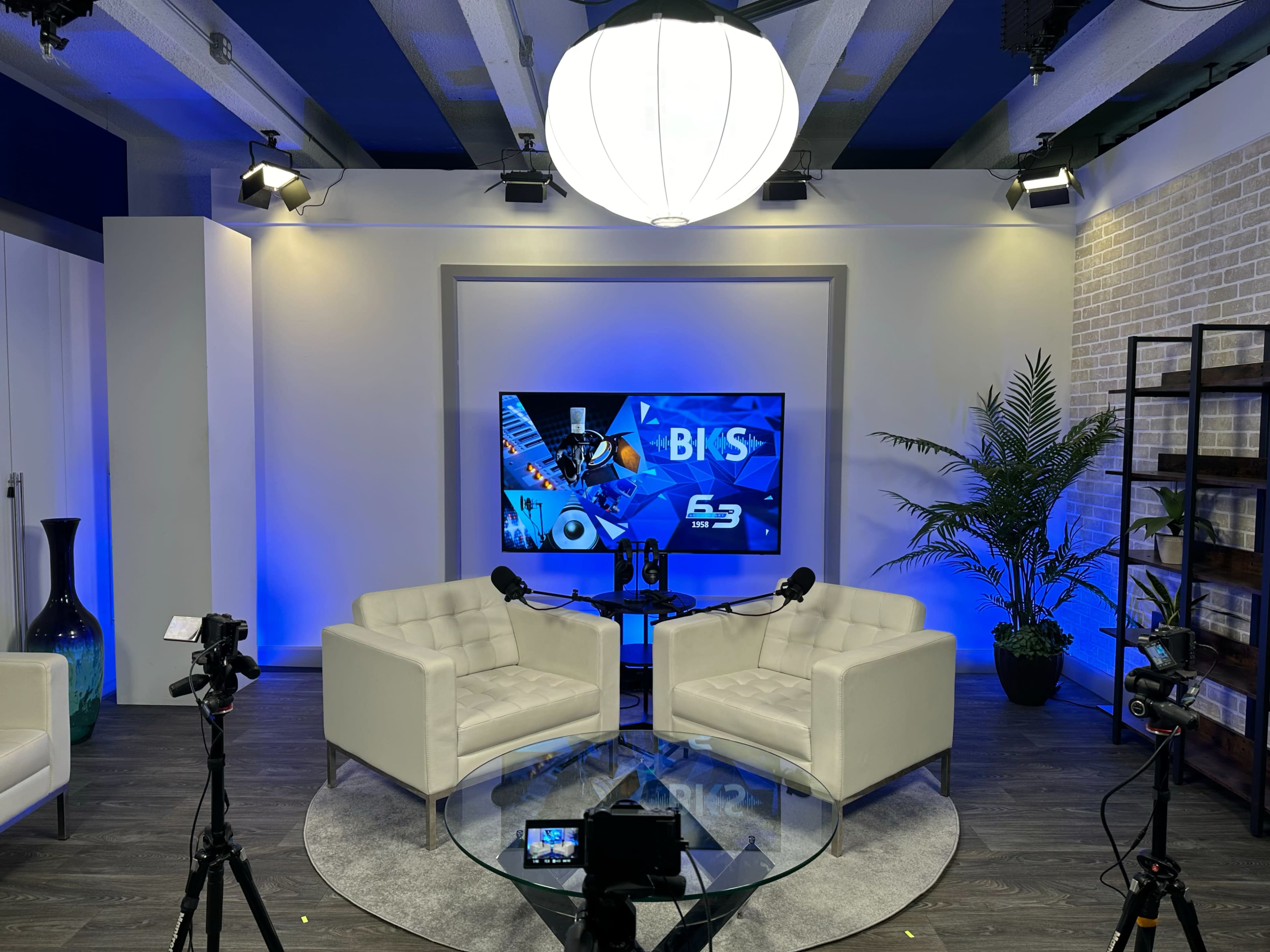 A modern studio setup with two white chairs facing a screen displaying graphics, surrounded by plants and cameras.