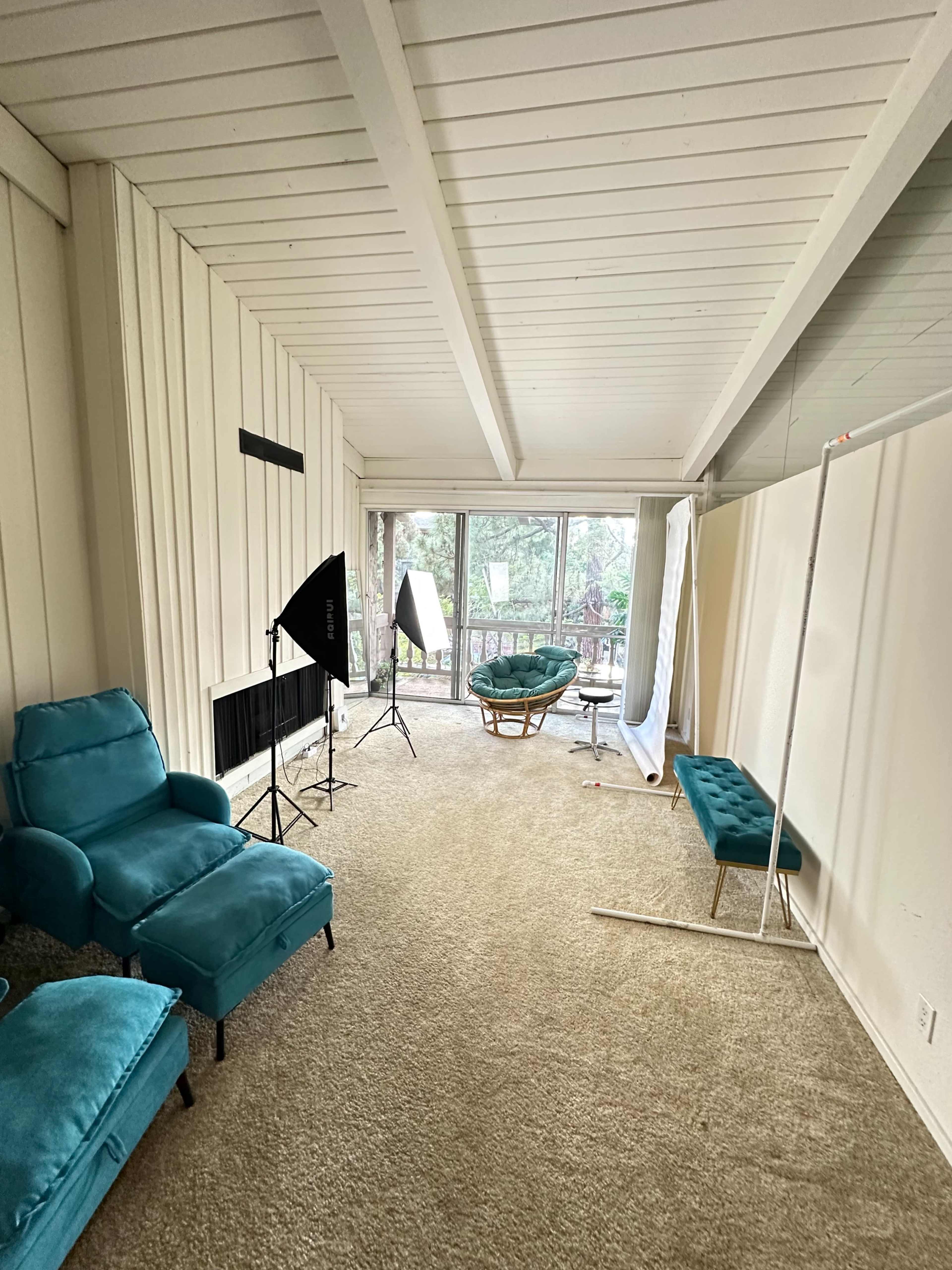 The image shows a spacious room with a light-colored carpet, teal armchairs, a green lounge chair, and several photography lighting equipment set up near large windows.