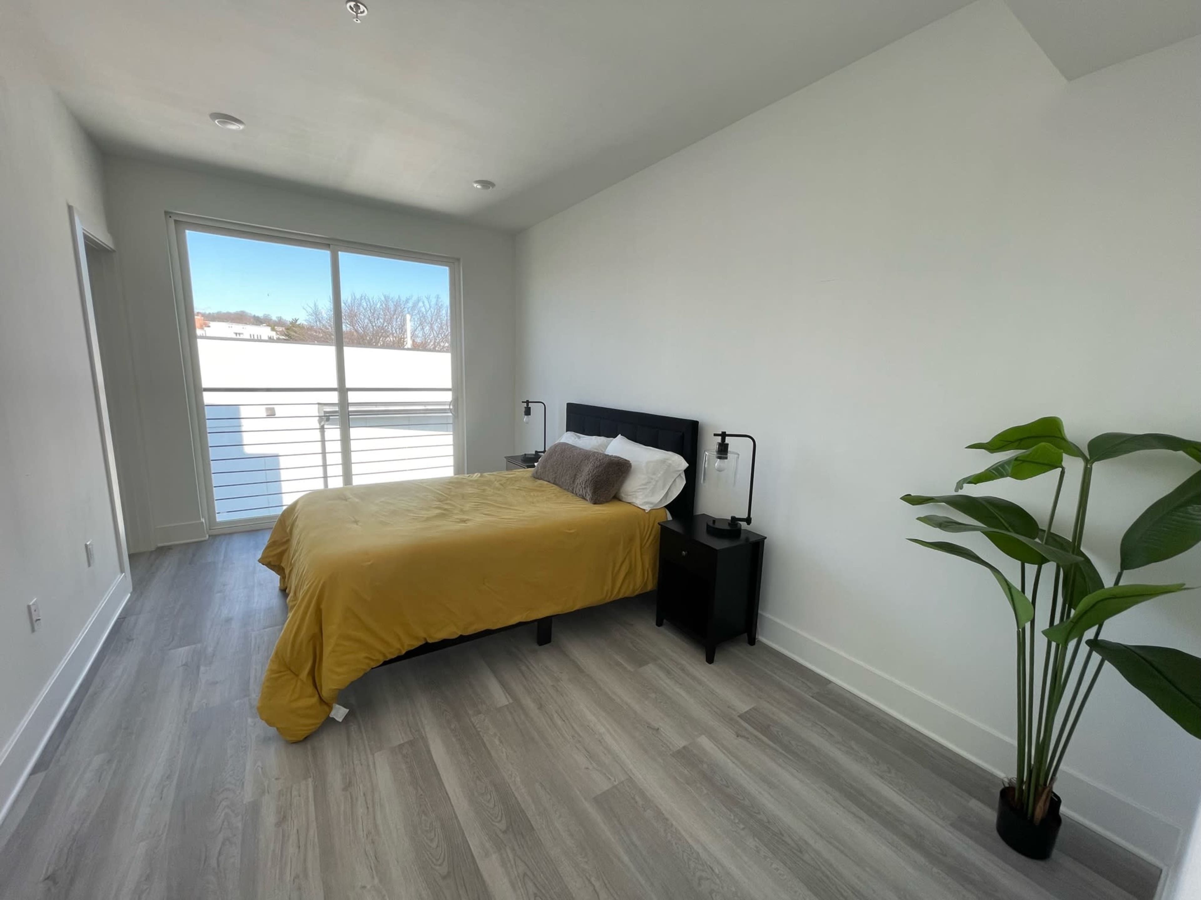 A minimalist bedroom with a yellow bedspread, a nightstand, and a potted plant near a large window.