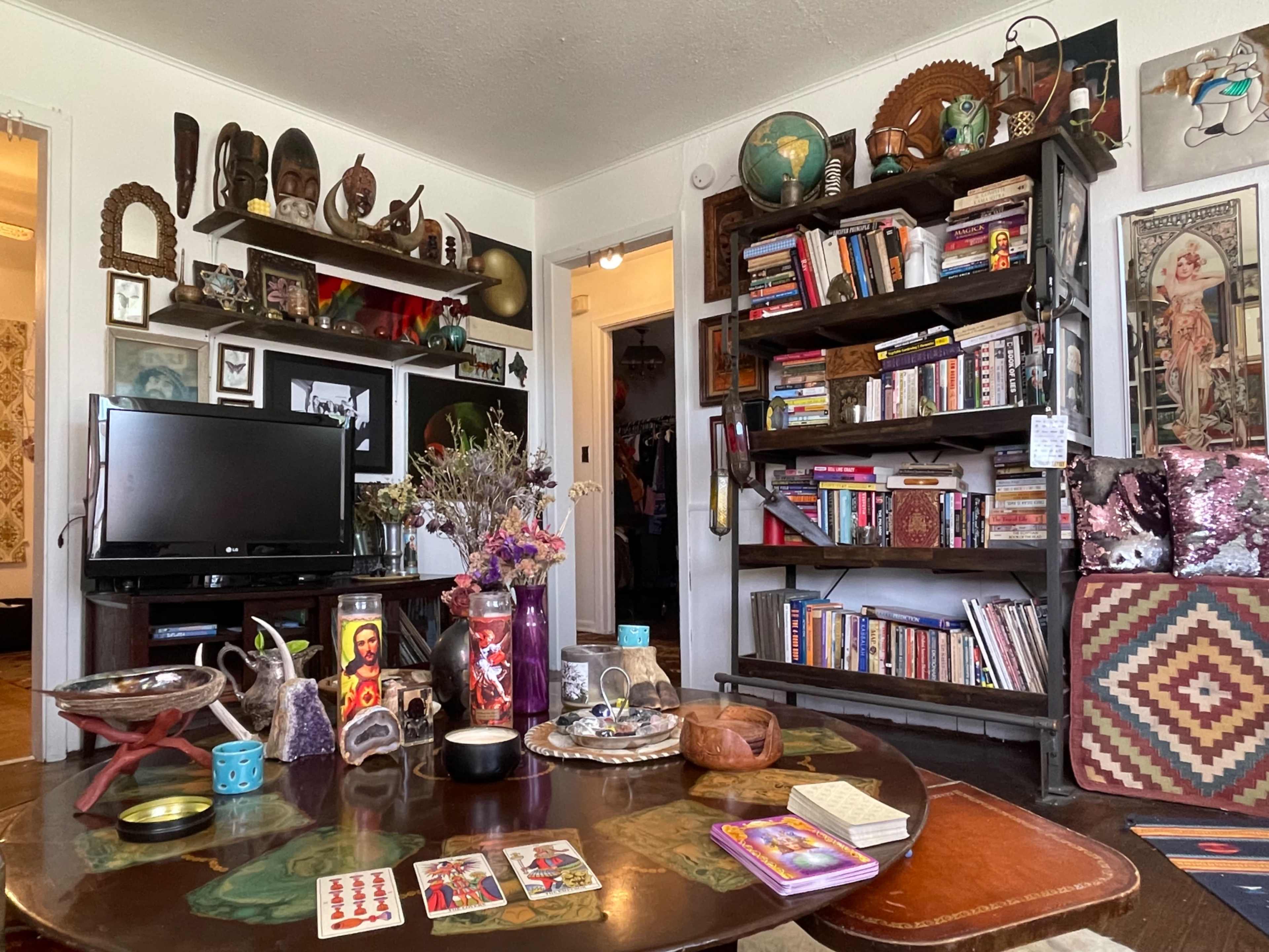 A cluttered living room with a wooden table at the center, surrounded by shelves filled with books, decorative items, and a television.