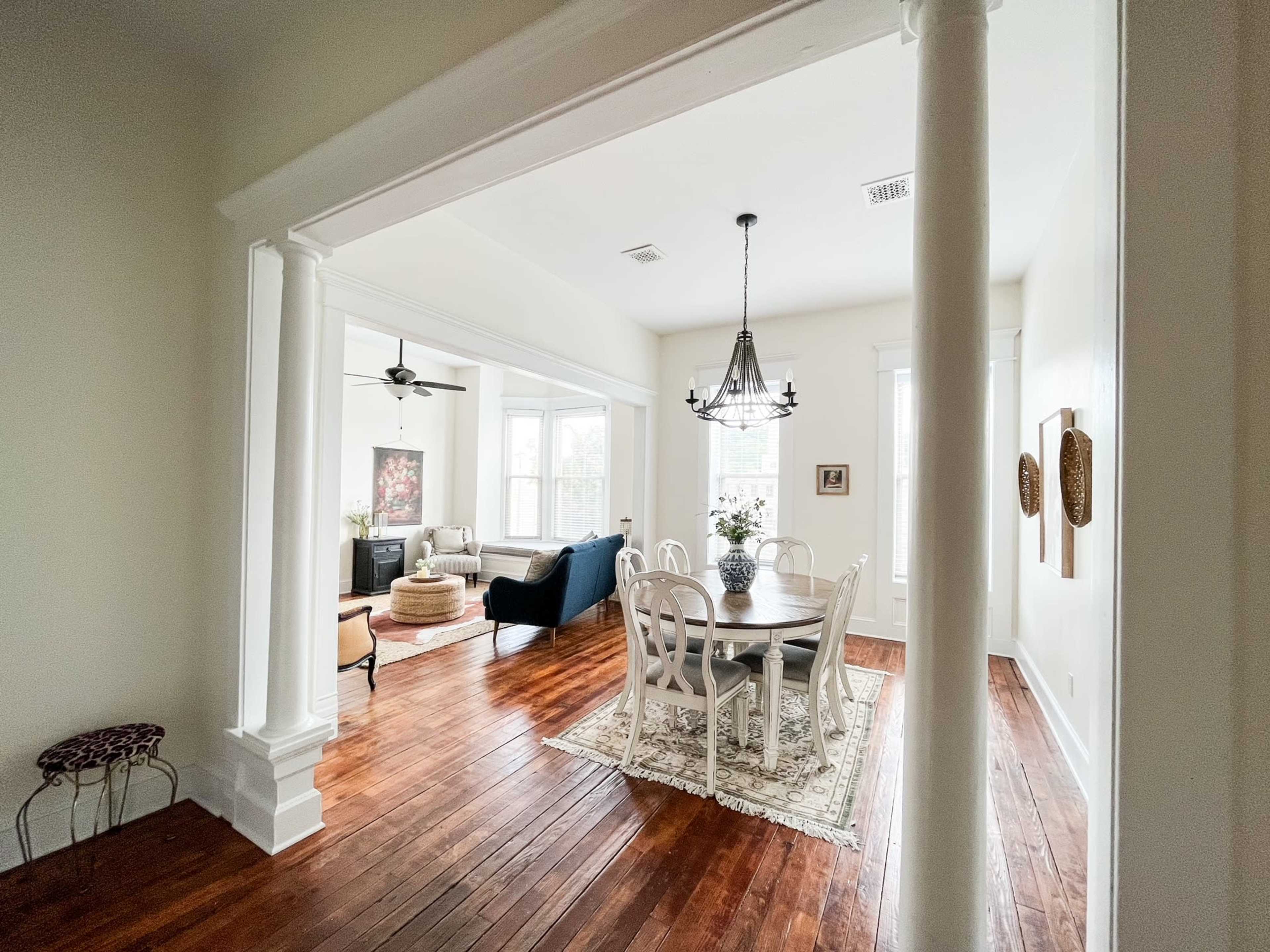 The image shows a room with a dining area featuring a round table and chairs, leading into a living space with a couch and large windows.