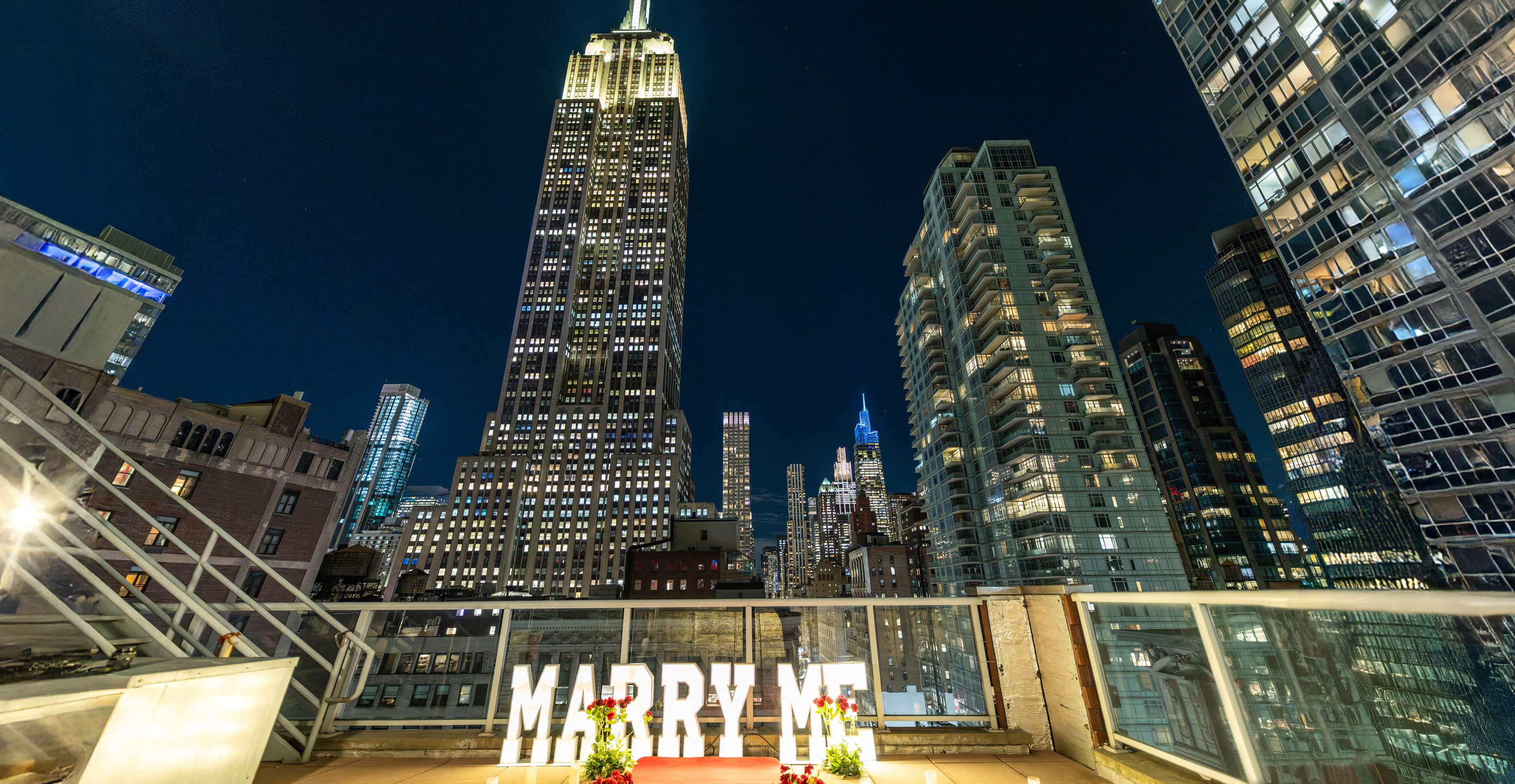 A romantic rooftop proposal scene features large light-up letters spelling "MARRY ME," with the Empire State Building illuminated in the background against a night skyline.