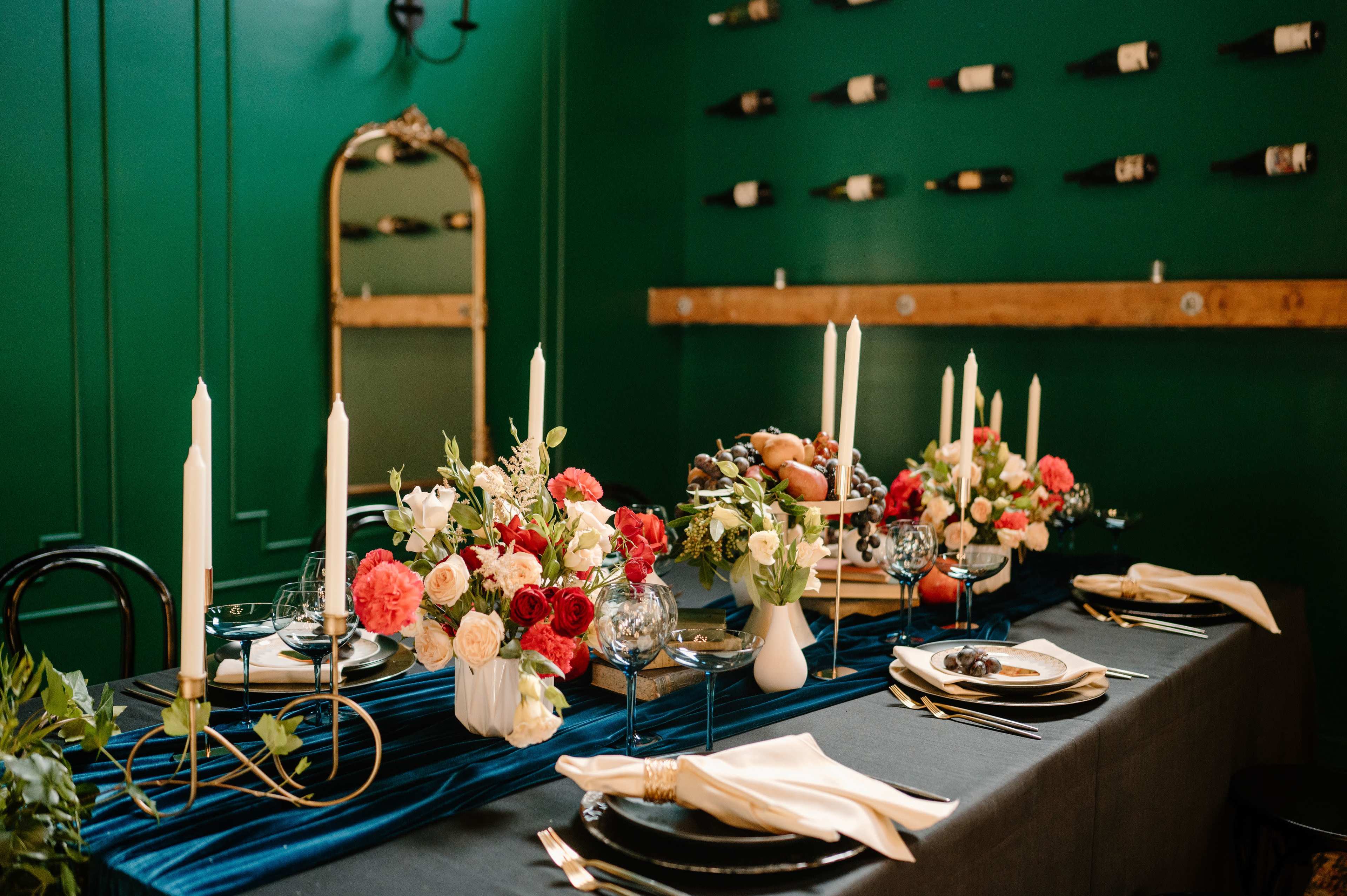A long dining table is set with floral arrangements, candles, and elegant dinnerware against a green wall decorated with wine bottles.