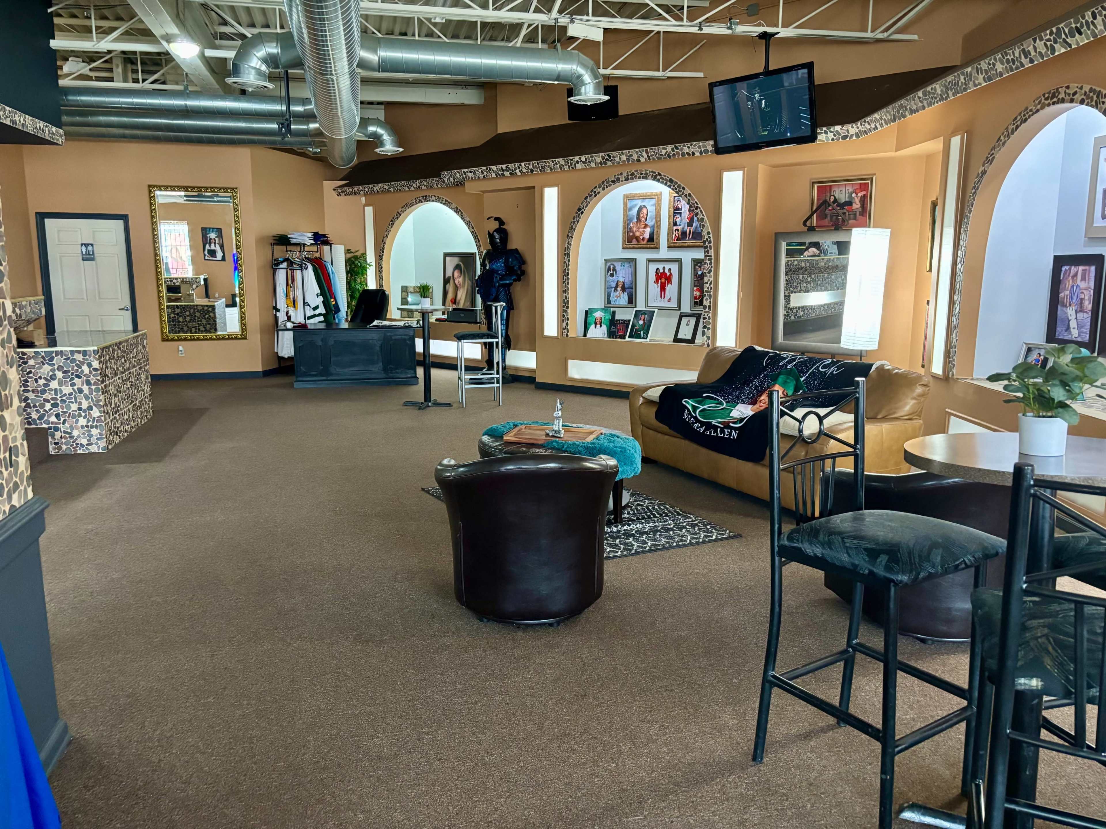 The image shows a spacious interior of a modern salon or studio, featuring a mix of seating areas, decorative mirrors, and a variety of displayed artwork on the walls.