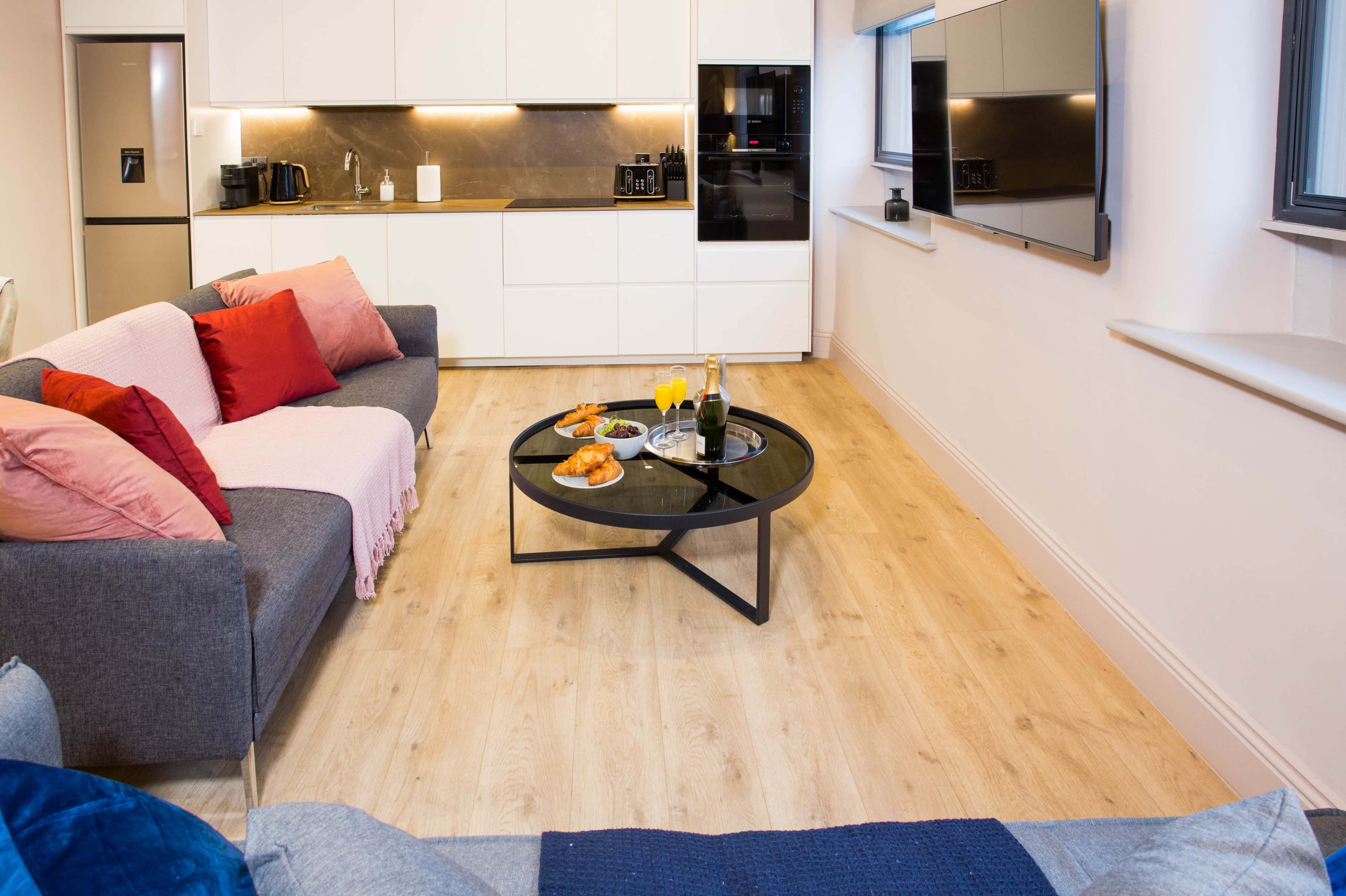 The image shows a modern living room with a gray sofa, a round black coffee table featuring snacks and drinks, and a sleek kitchen in the background.