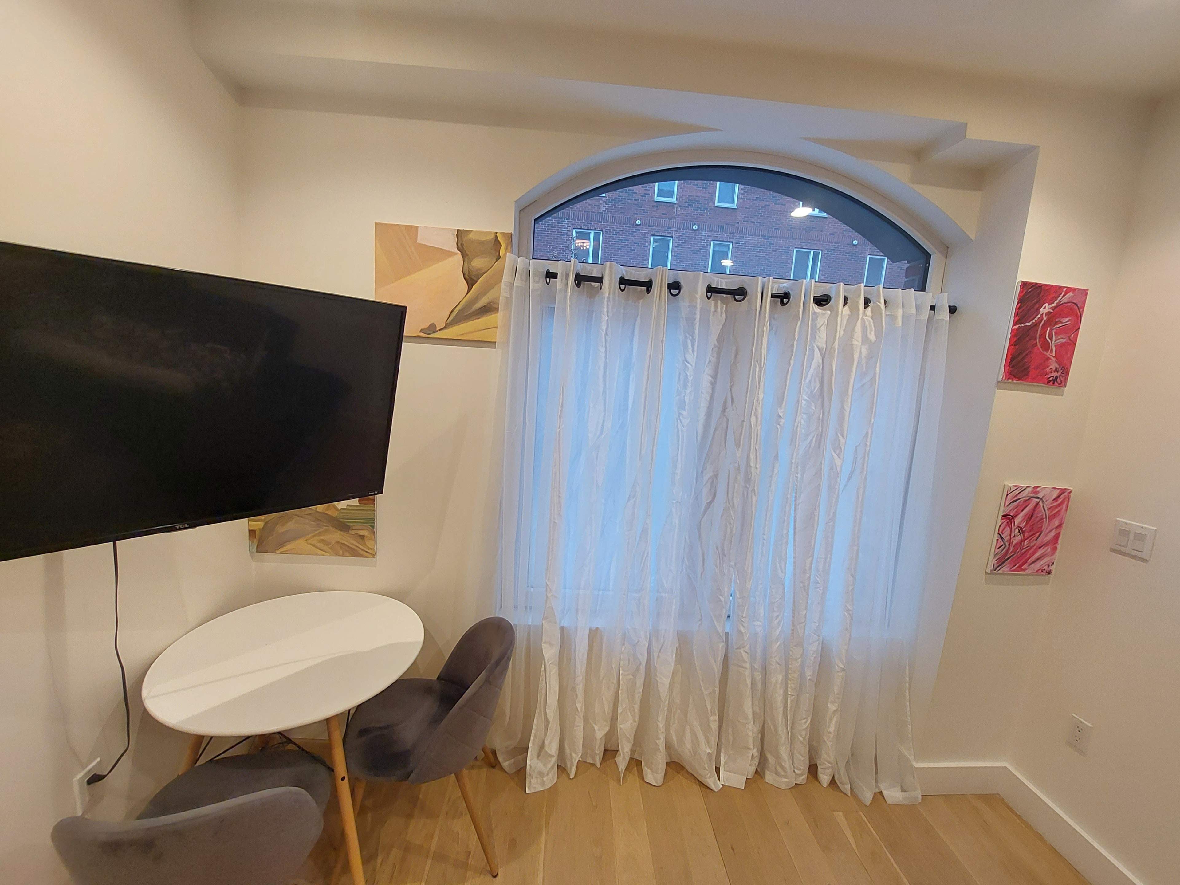 A small living area with a round white table and two gray chairs, a television mounted on the wall, and sheer white curtains framing a window with artwork displayed on the adjacent walls.