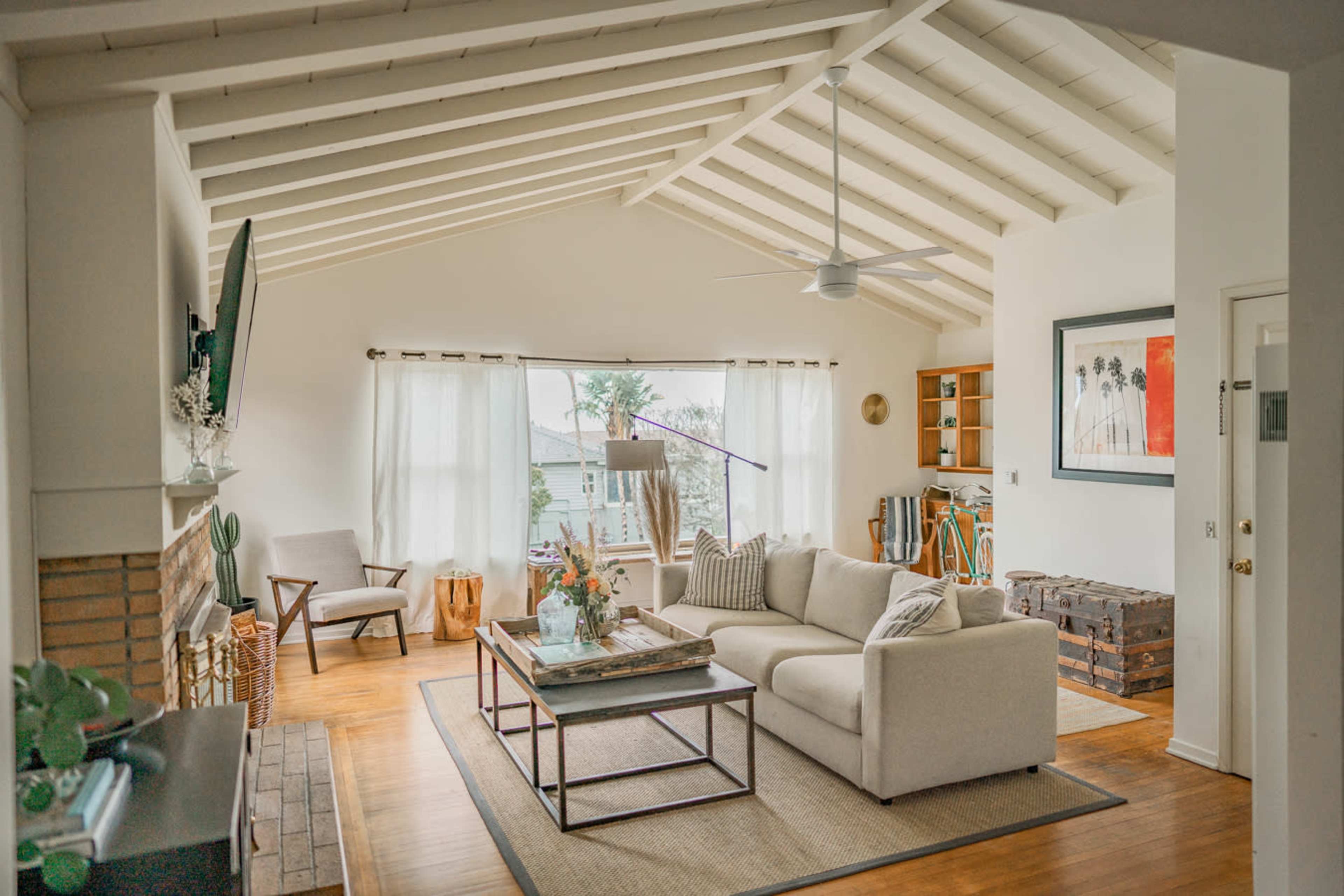 A spacious living room features a beige sectional sofa, large windows with sheer curtains, and a wooden coffee table, complemented by a ceiling fan and a fireplace.