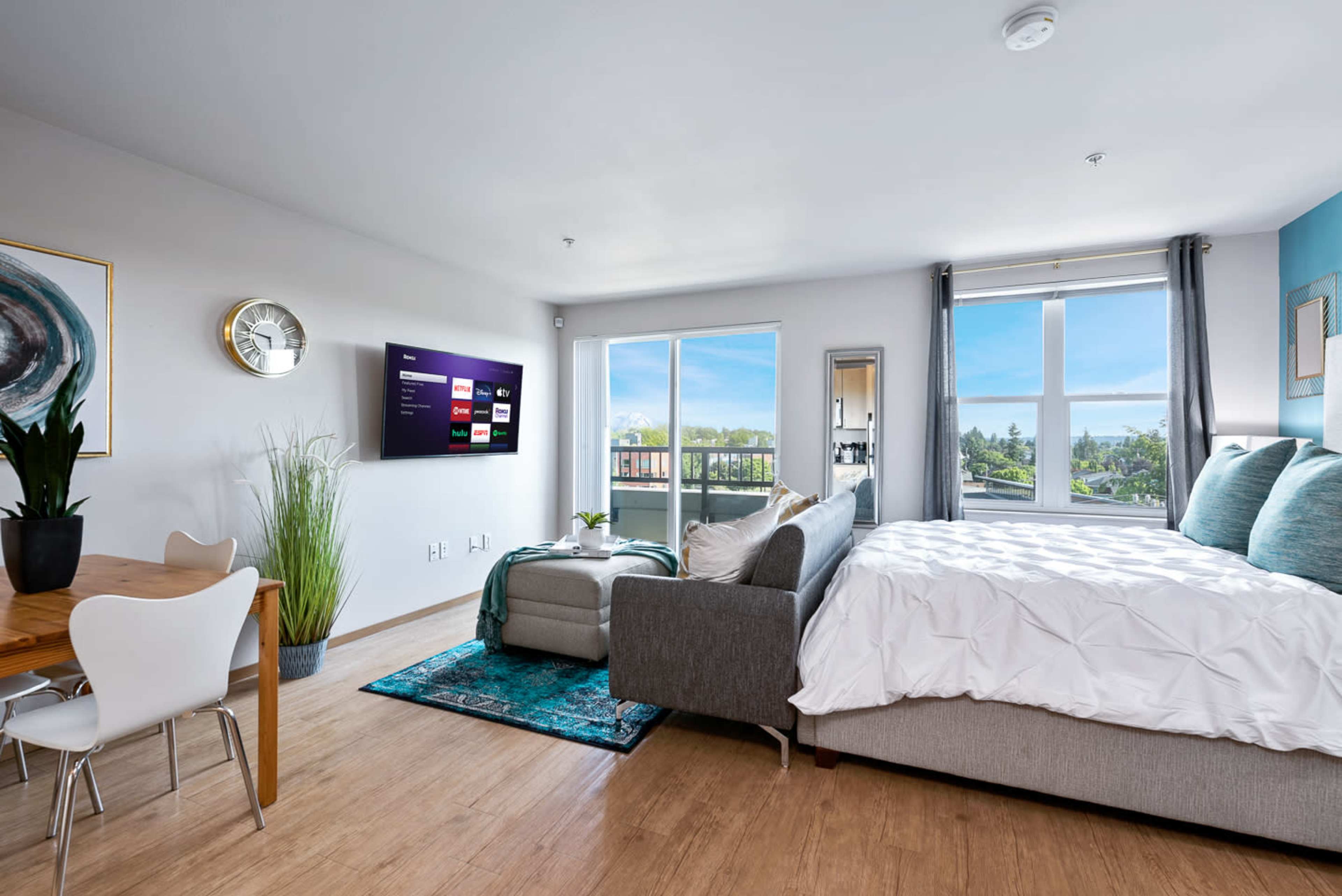 A modern studio apartment features a bed, a sofa, a dining table, and a wall-mounted TV with large windows displaying an outdoor view.