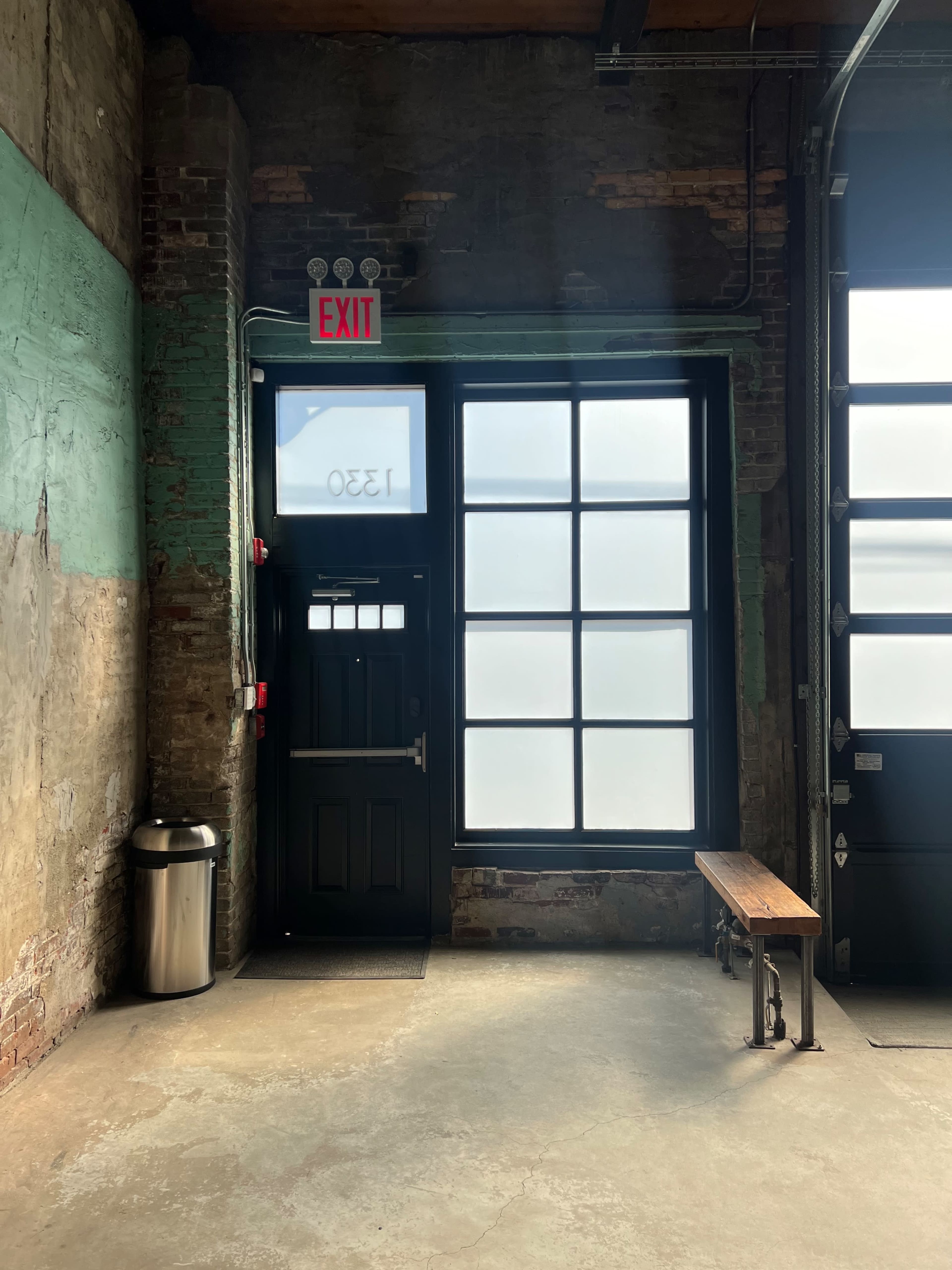 A room with exposed brick walls, featuring a large window and a black door with an exit sign, alongside a wooden bench and a trash can.