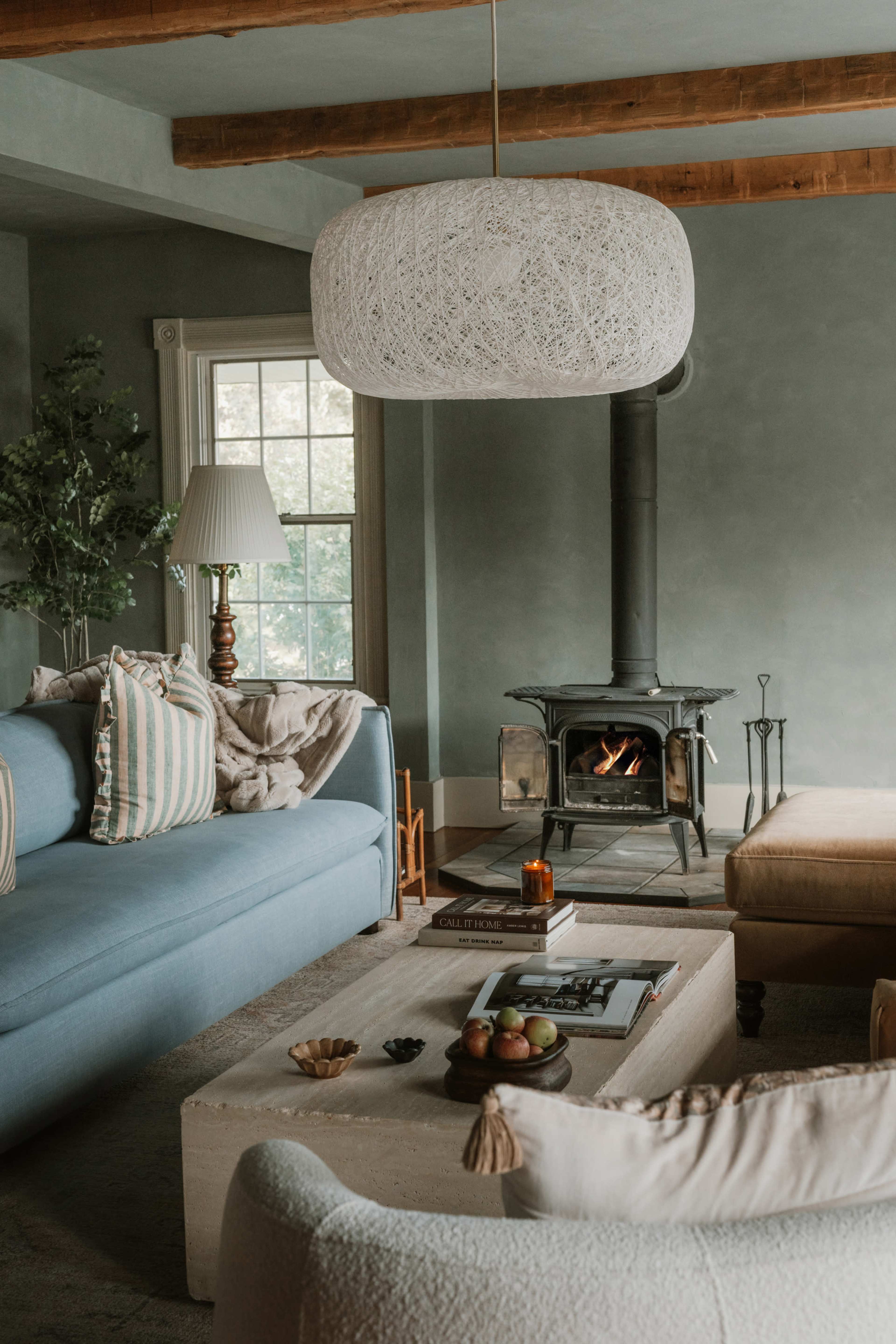 The image shows a cozy living room featuring a blue sofa, a light-colored coffee table, a wood-burning stove, and a large pendant light hanging from the ceiling.
