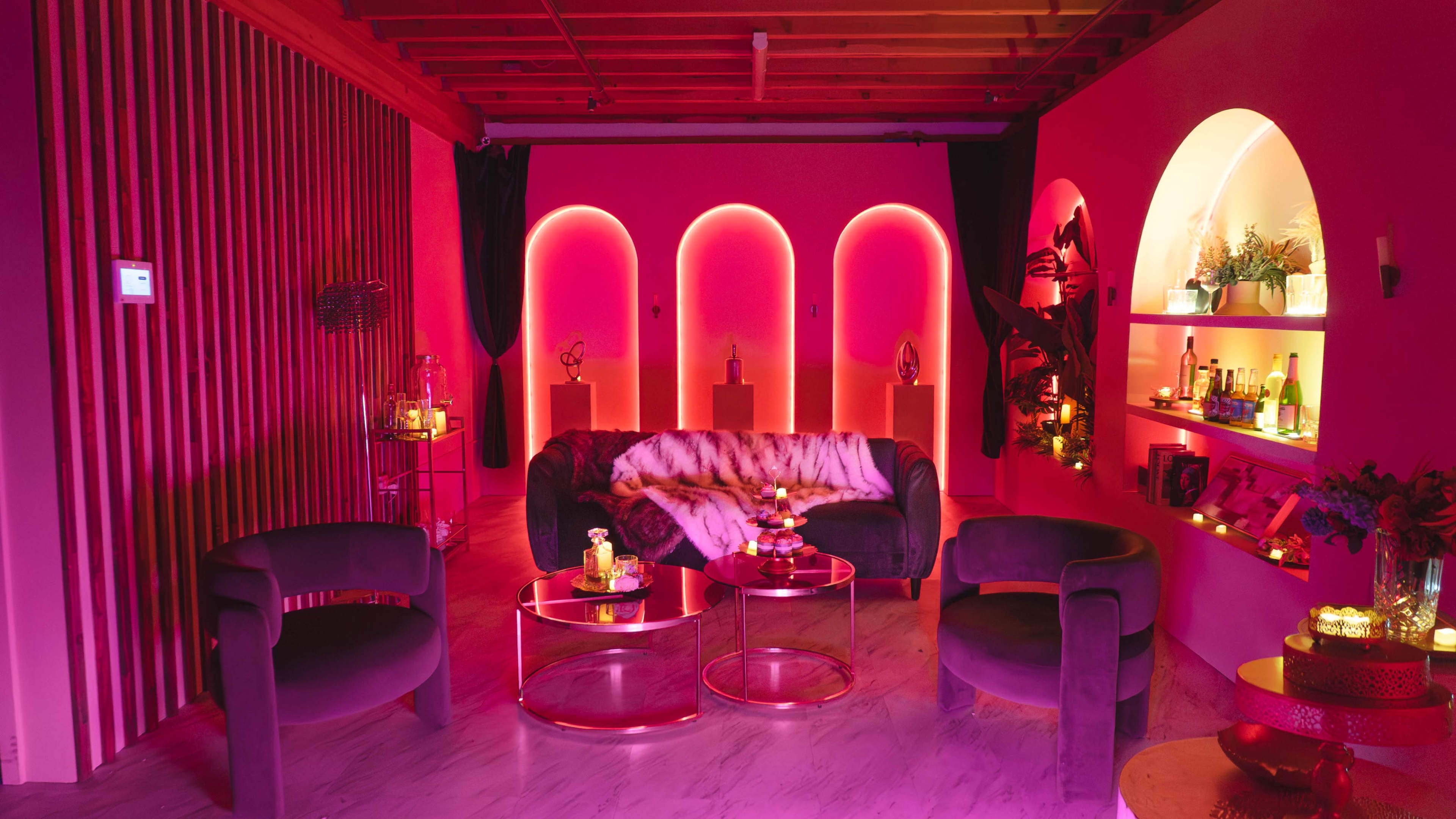 The image shows a stylish lounge area with pink walls, modern furniture, and decorative lighting creating a cozy atmosphere.