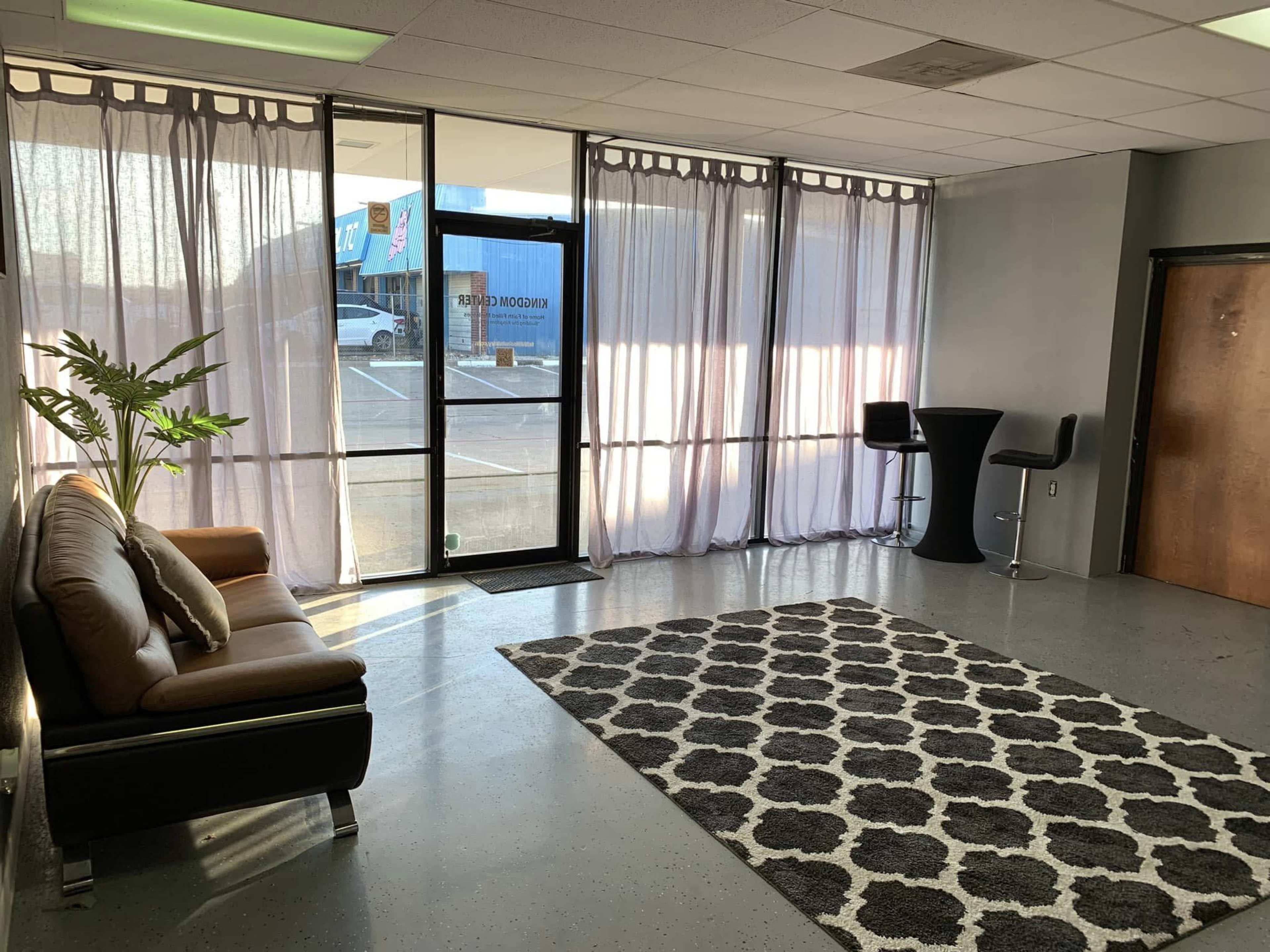 The image shows a minimalist waiting area furnished with a sofa, a round table, two bar stools, and a patterned rug, illuminated by natural light through large windows covered with sheer curtains.