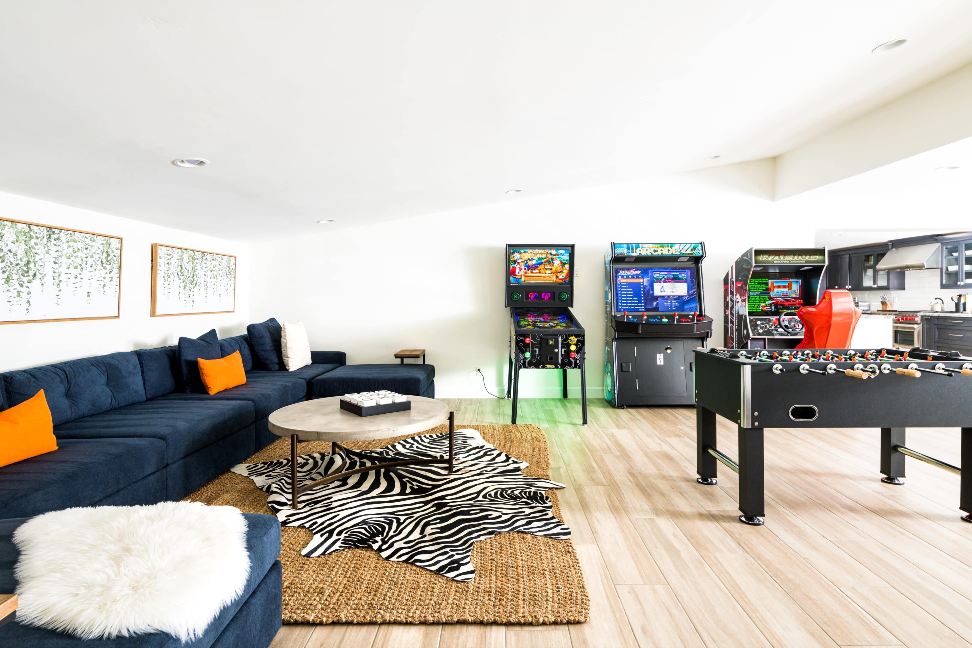 The image shows a modern game room featuring a dark blue sectional sofa, a coffee table on a zebra-patterned rug, and several arcade machines alongside a foosball table.