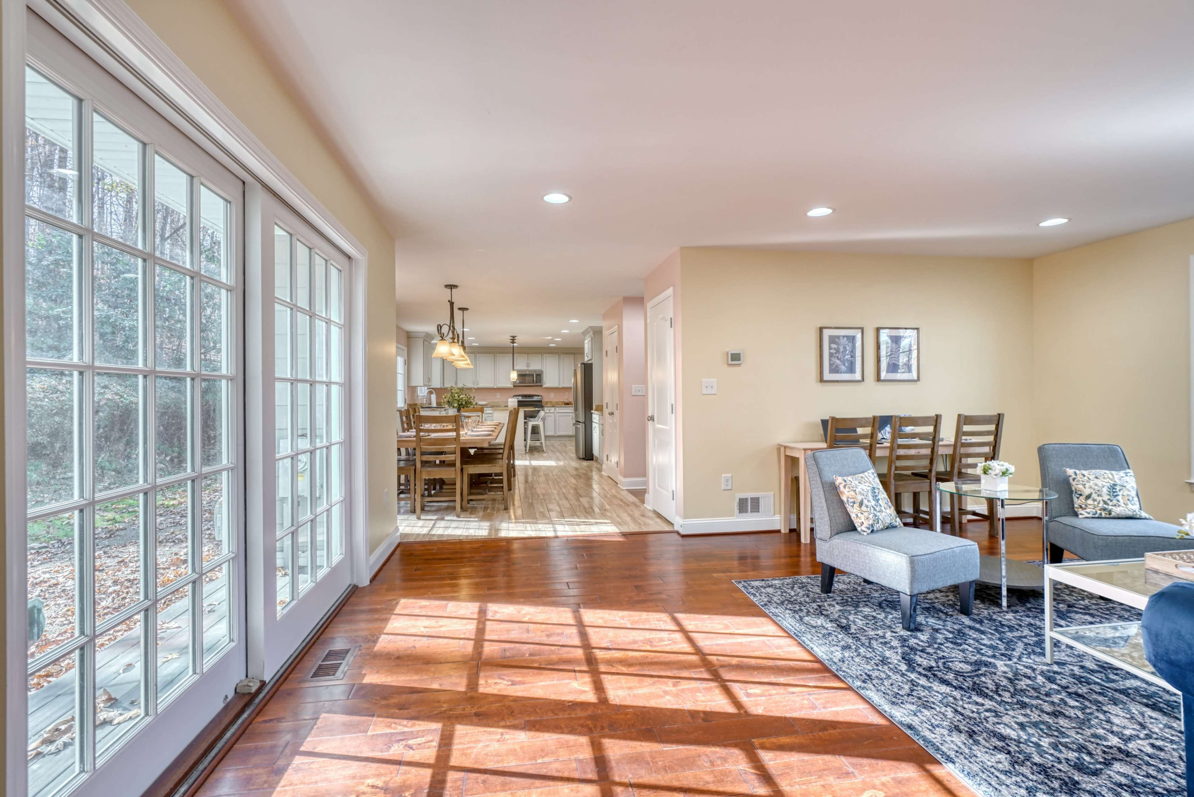A bright interior space featuring a living area with two gray chairs and a coffee table, leading to a dining area with wooden chairs and a kitchen visible in the background.