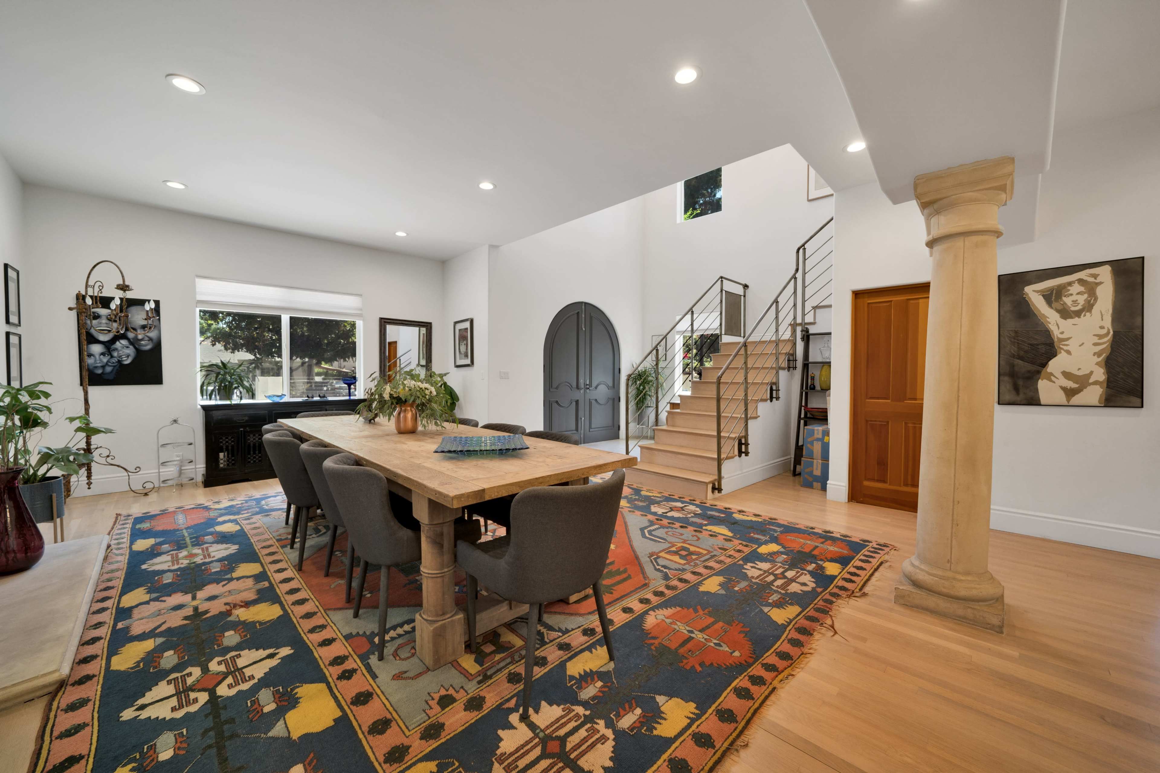 The image shows a spacious dining area with a large wooden table surrounded by gray chairs, adjacent to a staircase and a large window offering natural light.