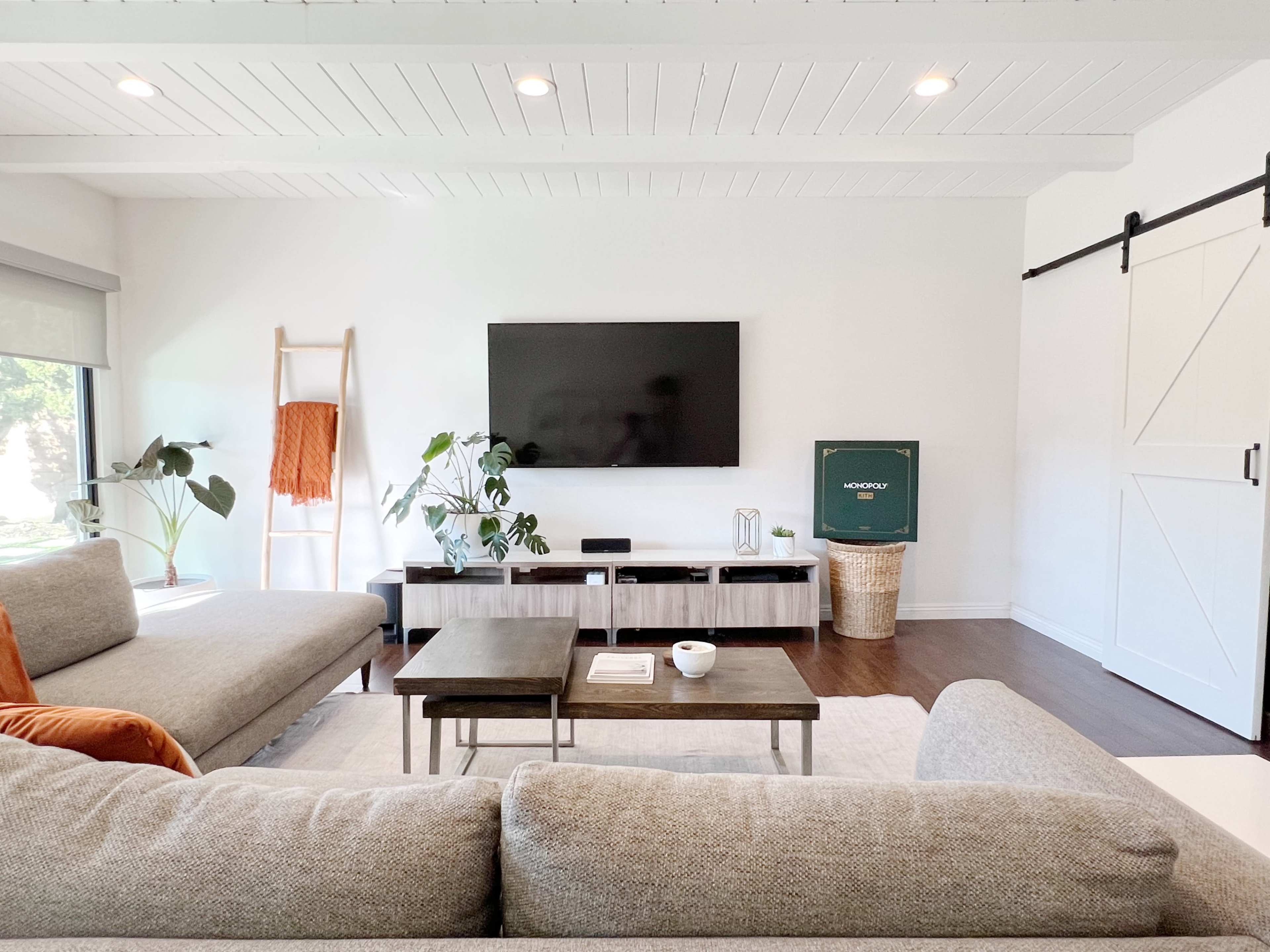 A modern living room features a gray sectional sofa, two coffee tables, a large TV mounted on the wall, and a sliding door leading to a bright space filled with plants.