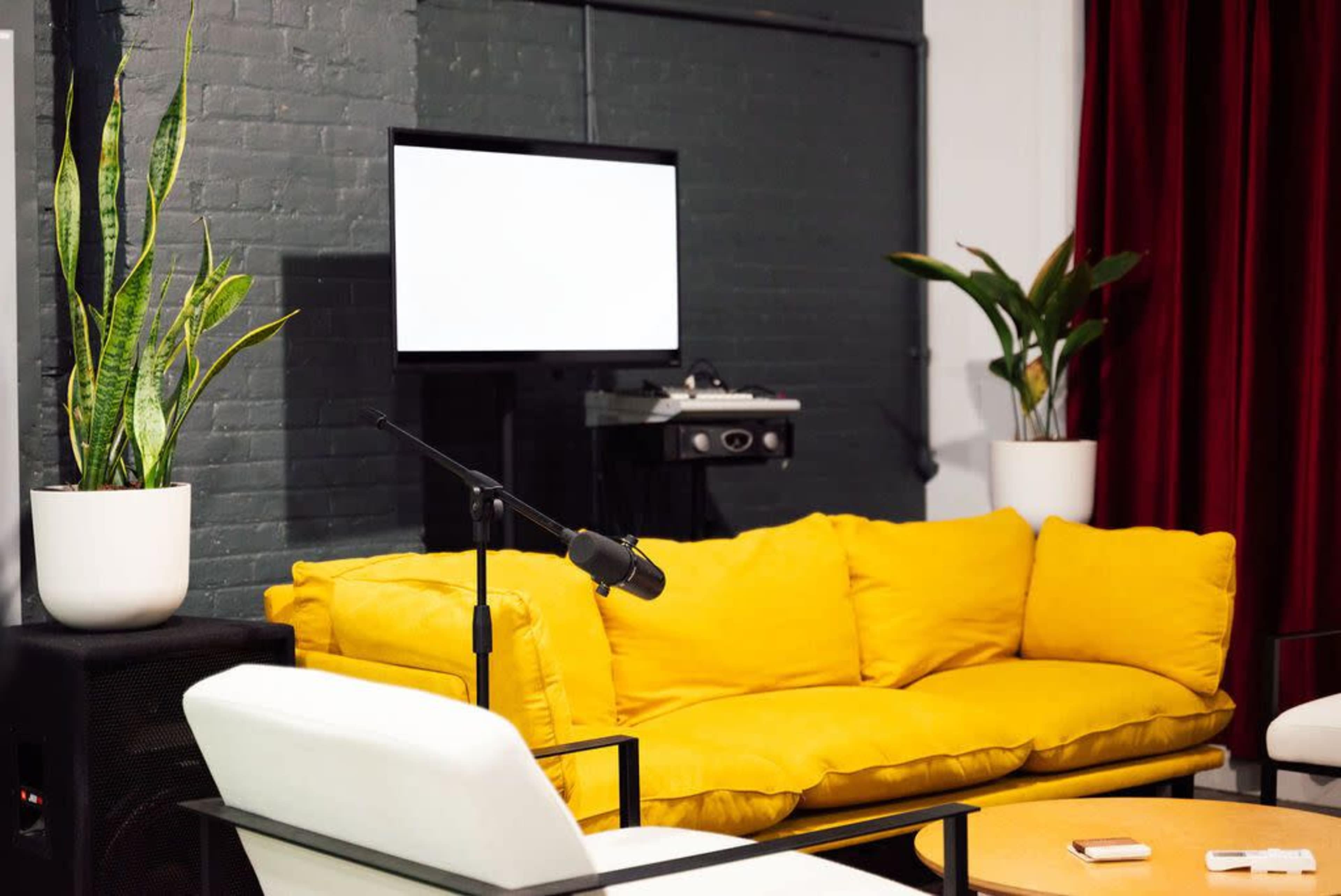 A modern living room with a yellow couch, a white chair, a television screen on the wall, and decorative plants in white pots.