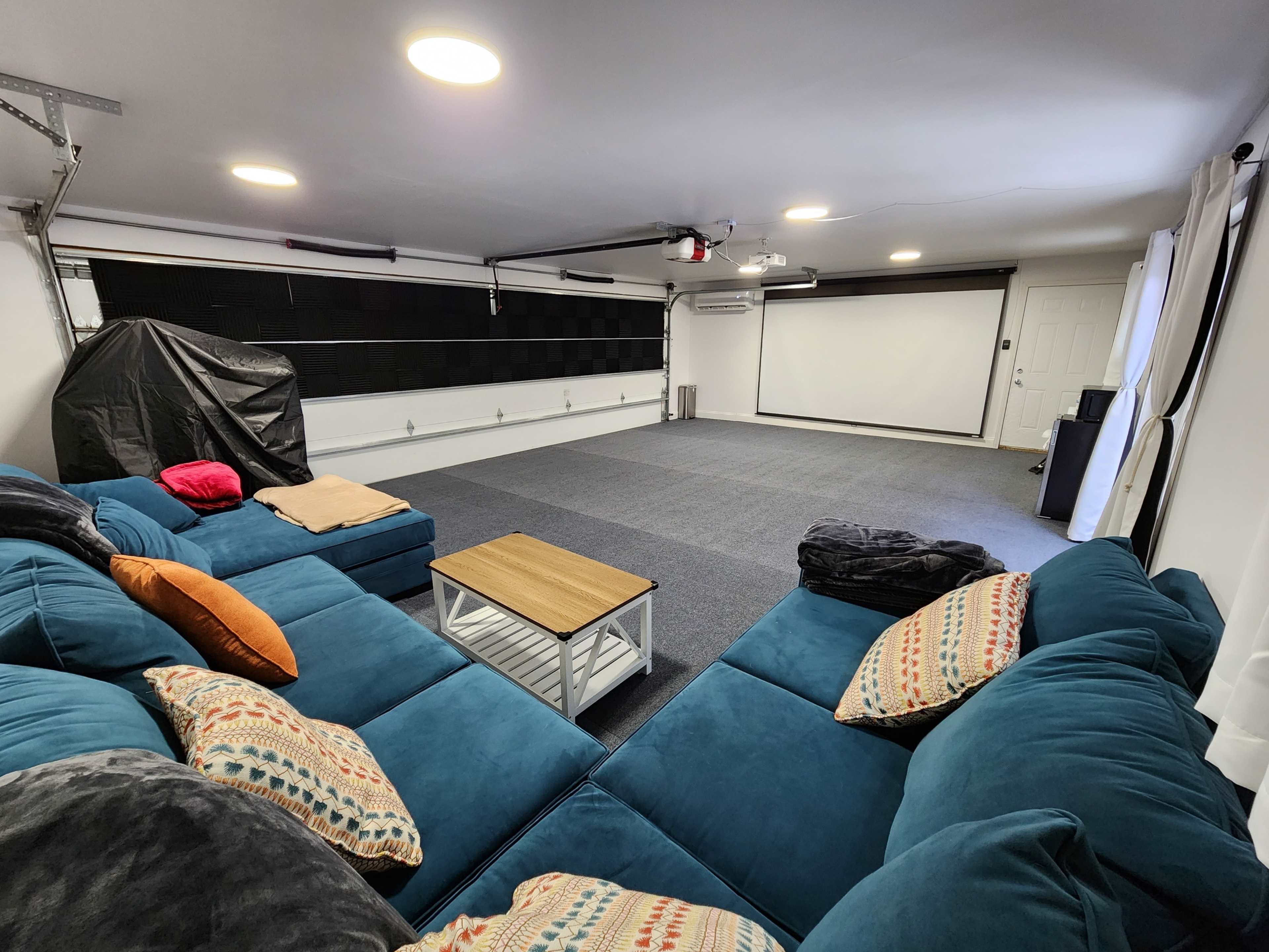 A spacious basement room featuring a large sectional couch, a coffee table, and a projection screen at one end.