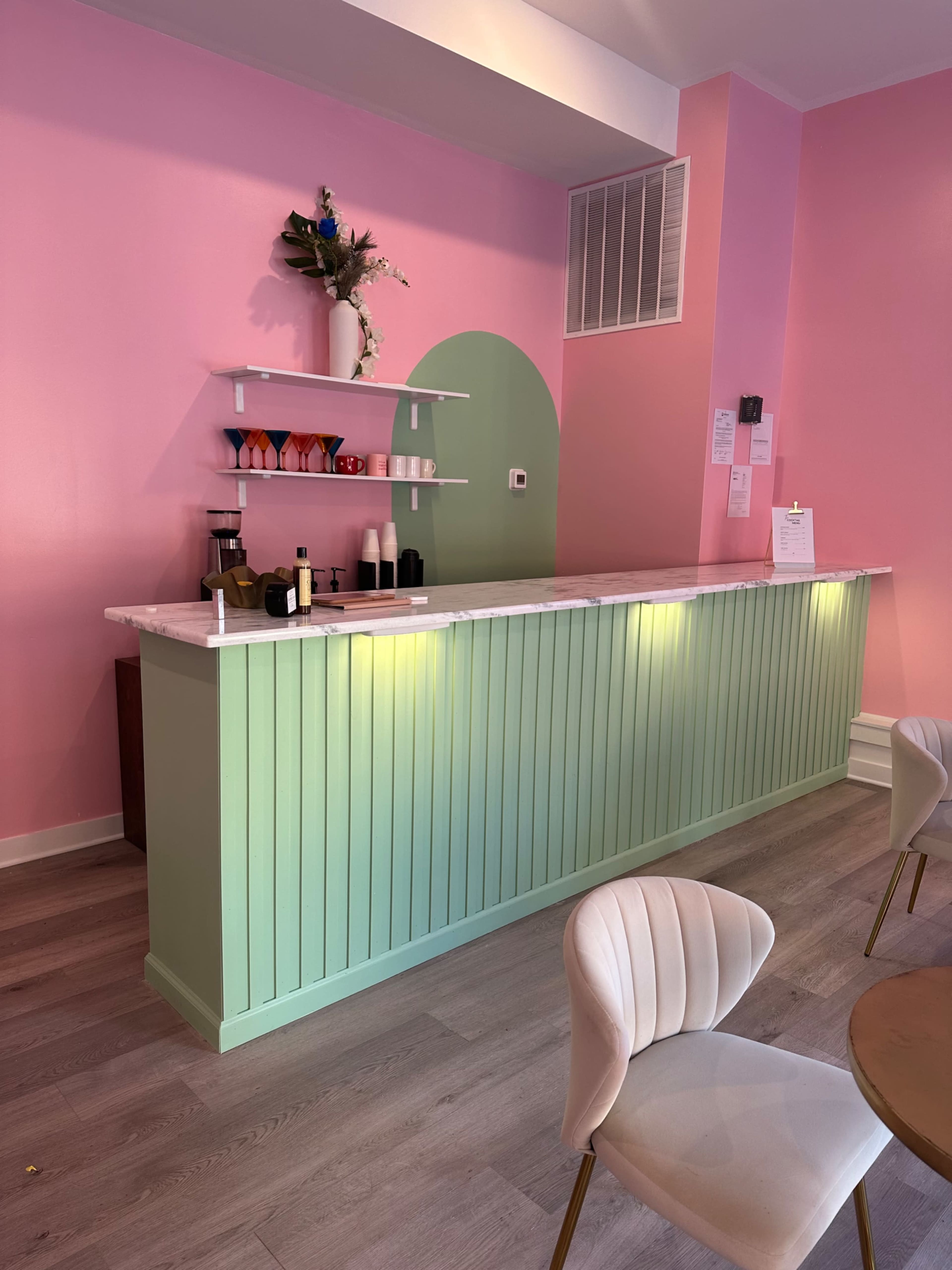 A brightly colored cafe interior with a green countertop and pink walls, featuring shelves with various cups and decorative items.
