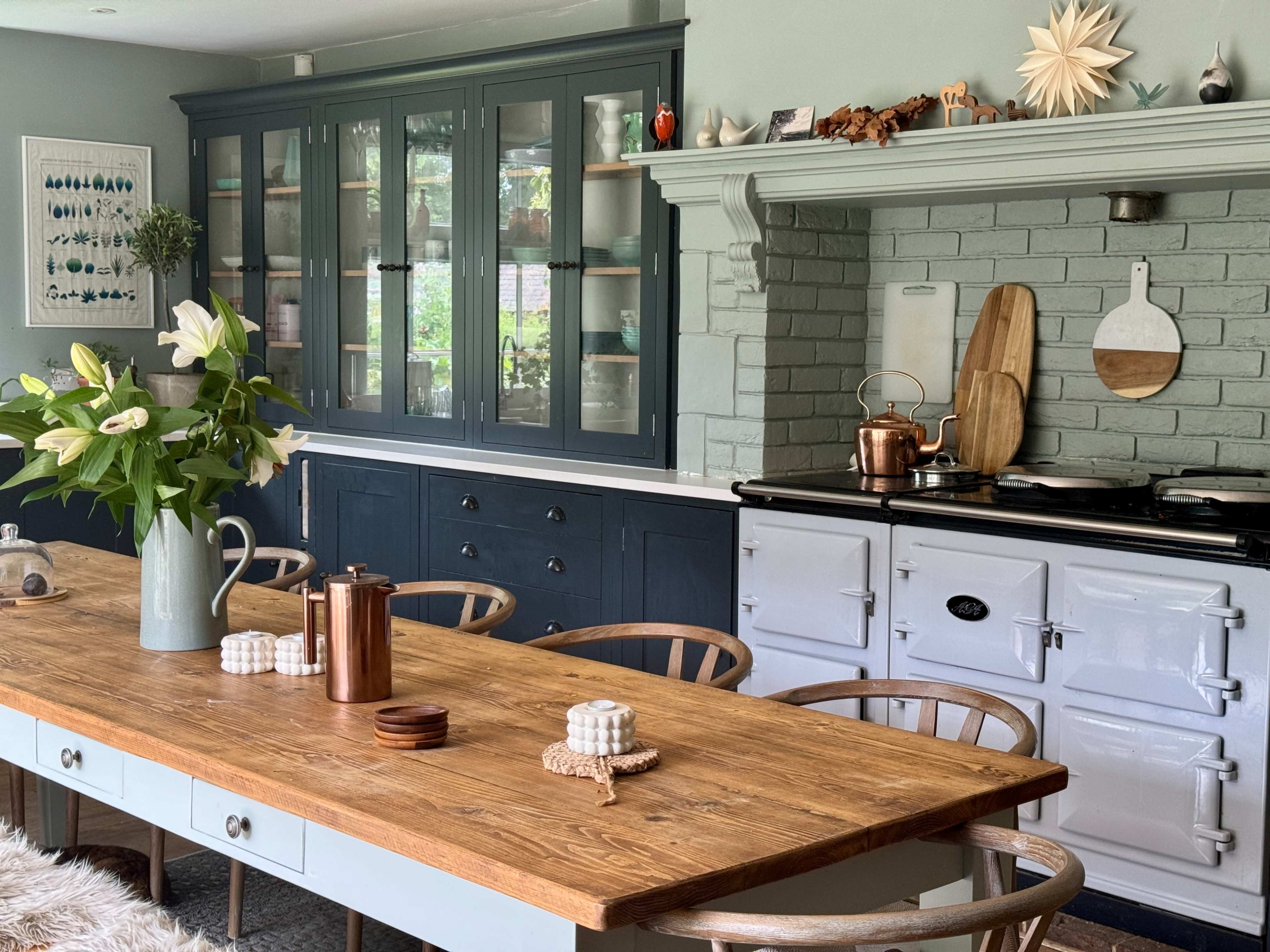 A modern kitchen featuring a large wooden dining table surrounded by chairs, with a blue-gray cabinet and an Aga-style stove against a green brick wall.