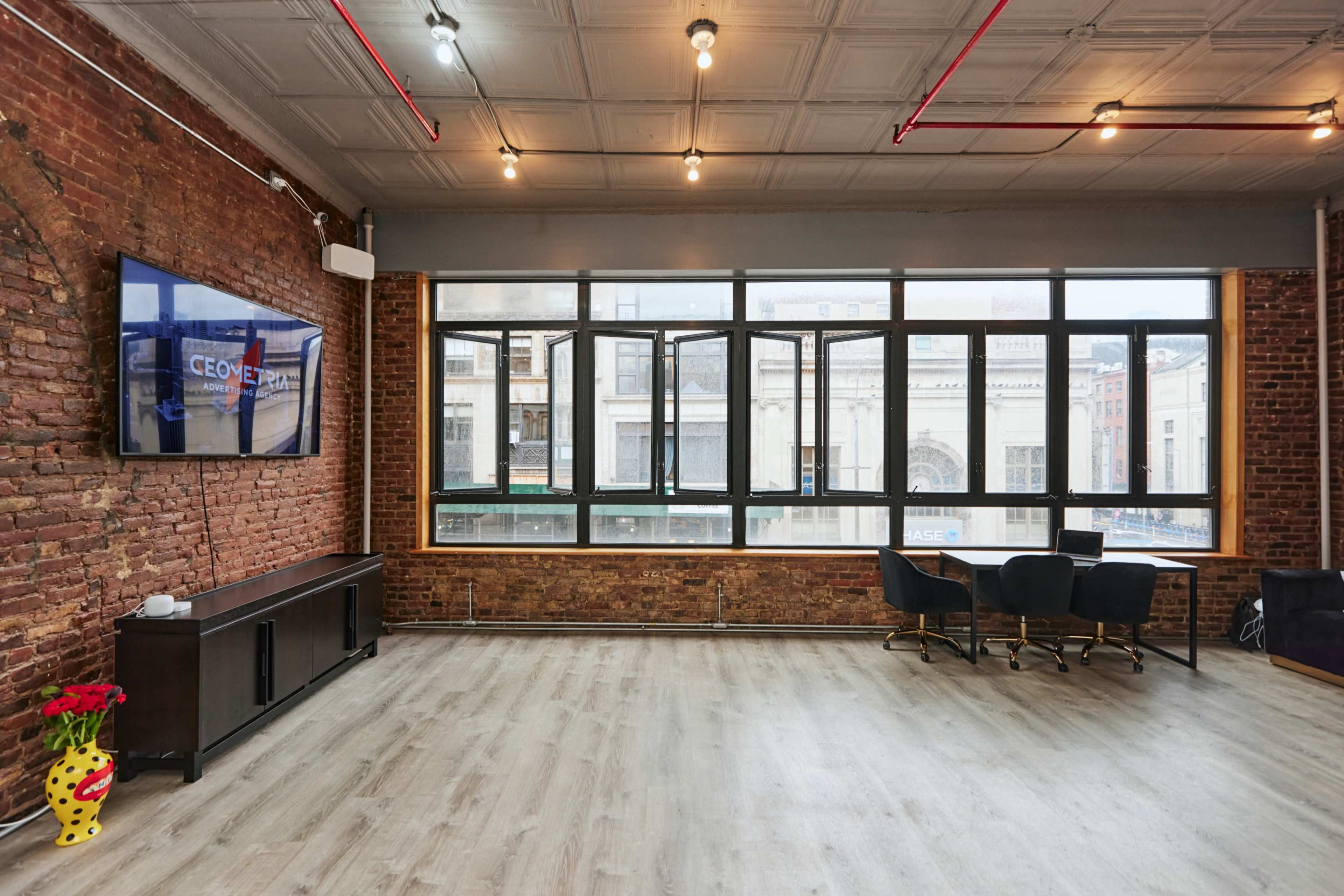A spacious room with exposed brick walls, large windows allowing natural light, a television on the wall, and a seating area with black chairs and a small table.