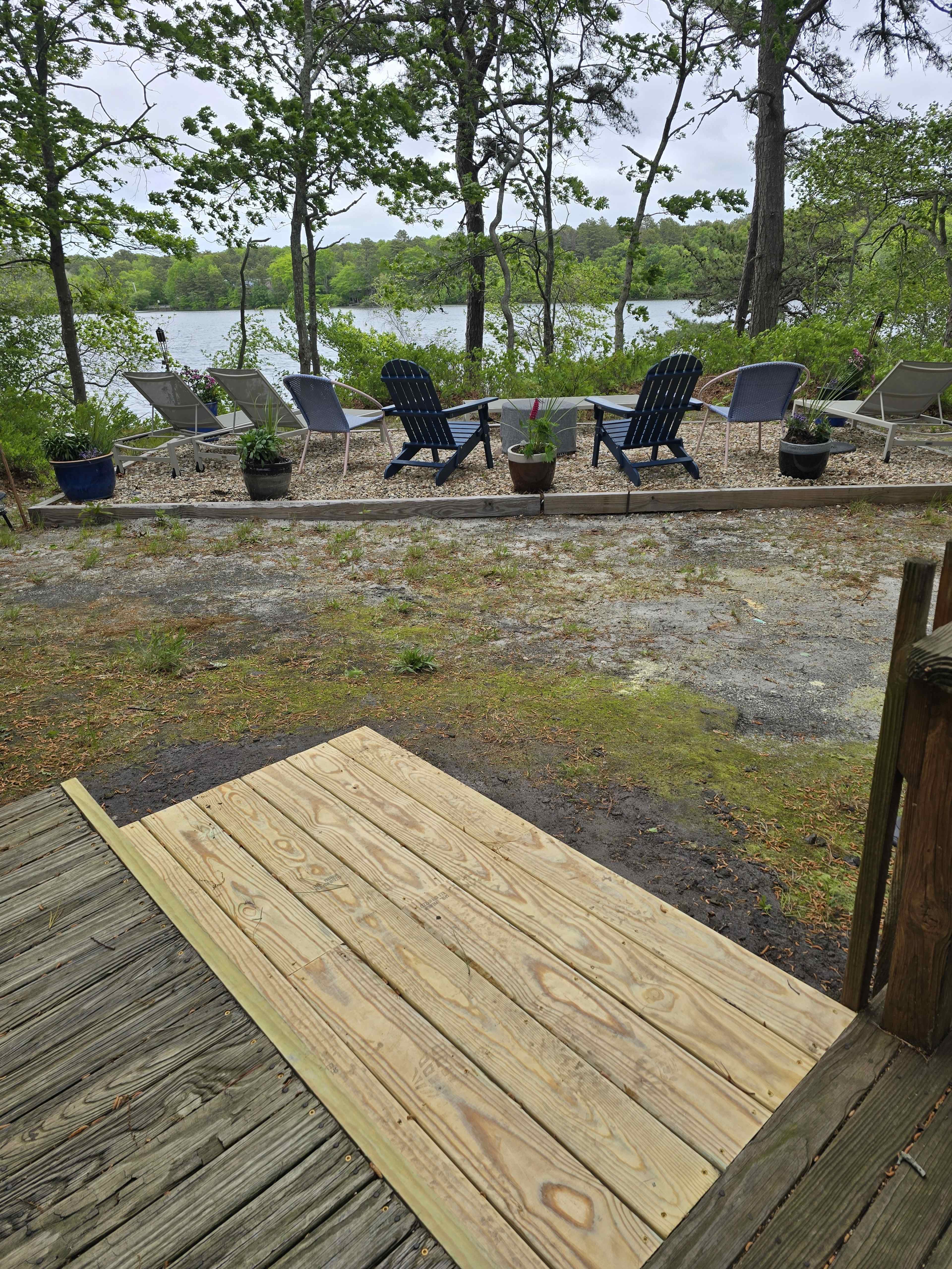 A wooden ramp leads down from a deck to a gravel area with several blue lounge chairs facing a body of water surrounded by trees.