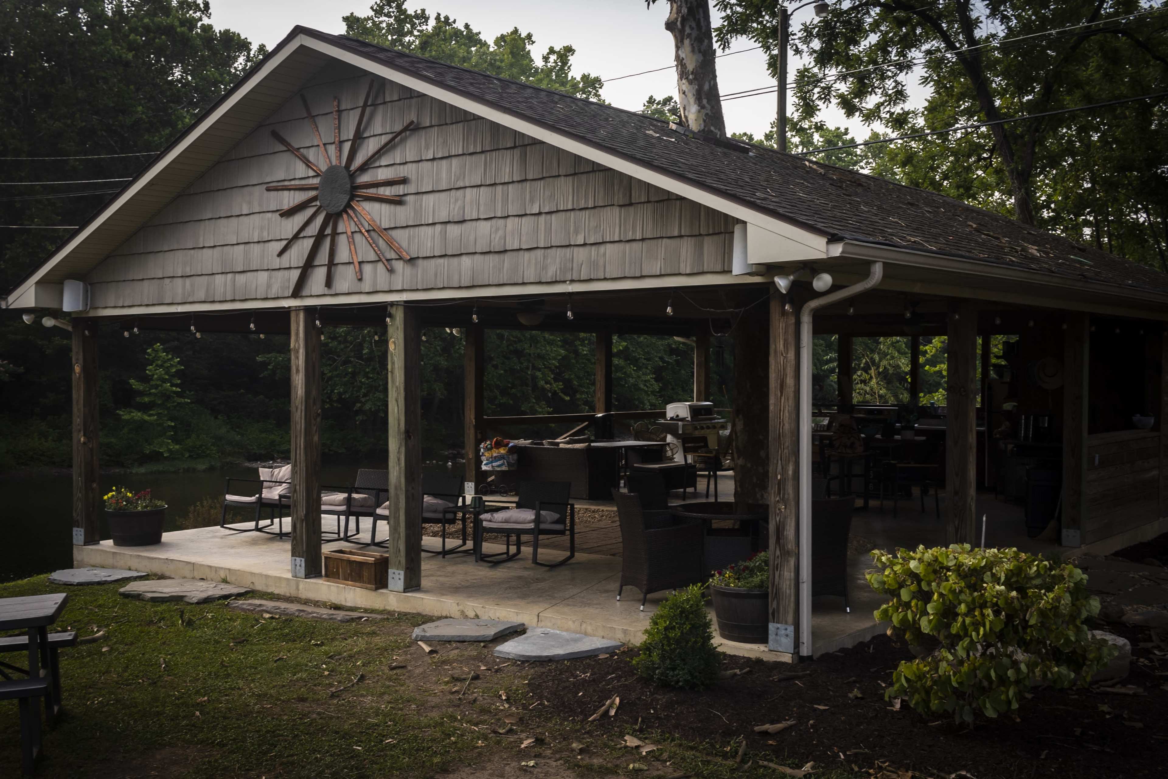 A wooden pavilion with a stone patio, featuring seating, a grill, and a large sunburst decoration on the wall, situated near a body of water surrounded by trees.